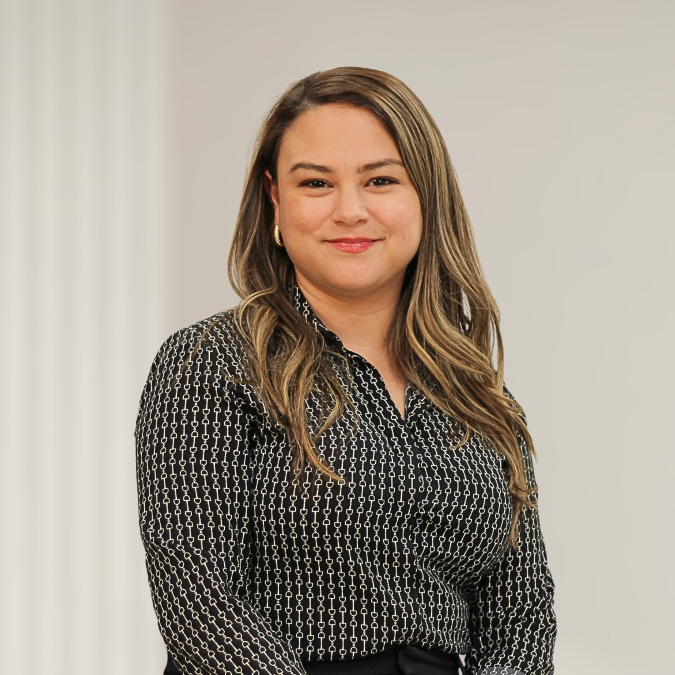 A young woman with light brown hair, wearing a navy blazer and white top, is smiling at the camera in front of wooden wall decor.