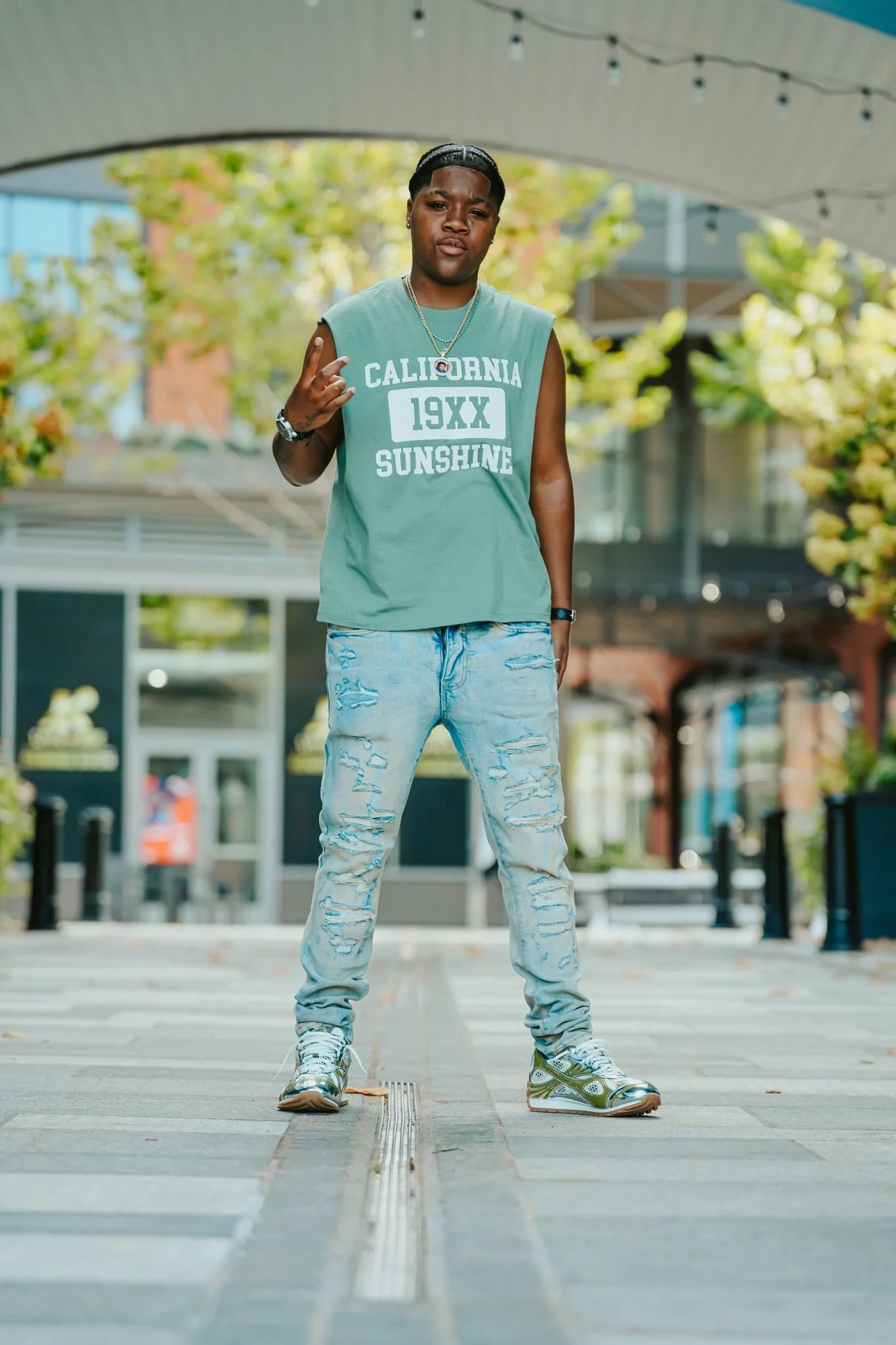 A young womanin a sleeveless green t-shirt with 'California 19XX Sunshine' written on it, distressed light blue jeans, and sneakers standing on a city sidewalk during daytime, making a peace sign with his right hand.