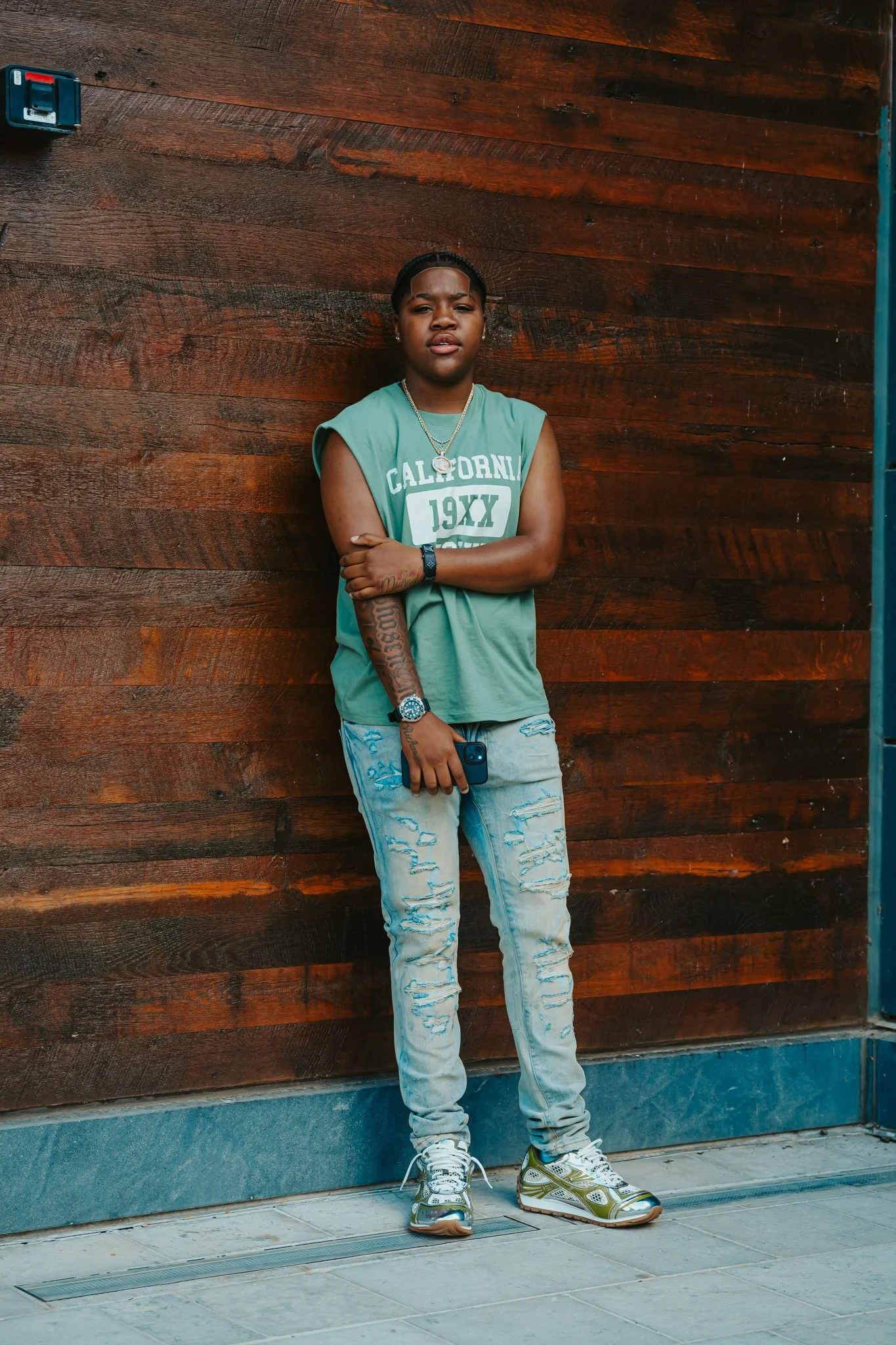 Young woman standing against a wooden wall, wearing a sleeveless green California t-shirt, distressed jeans, and gold sneakers, holding a phone in his right hand.