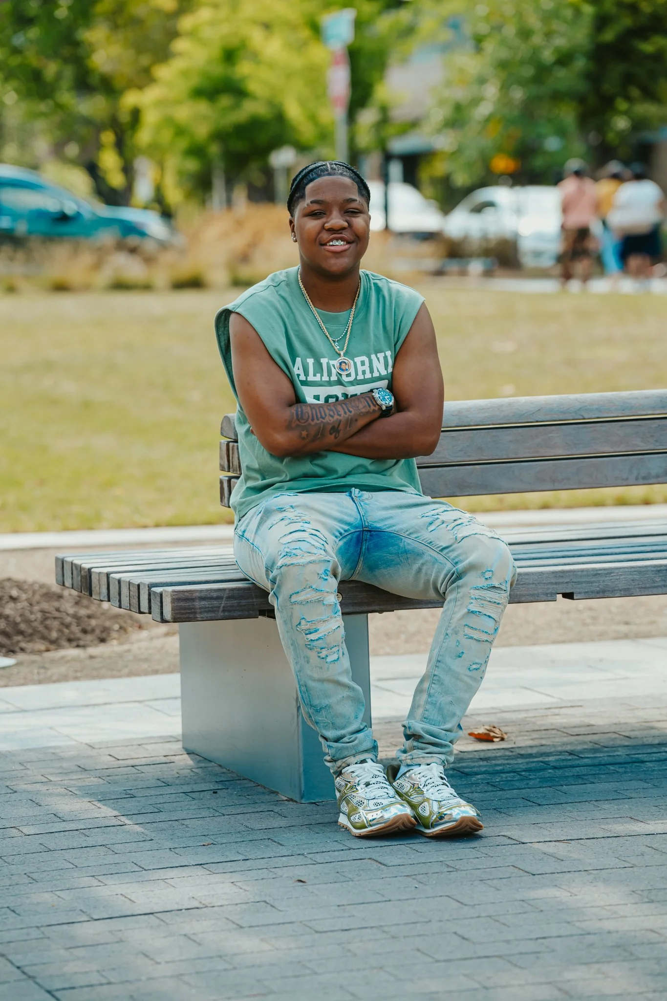 A young man with dark skin and short black hair, sitting on a wooden bench outdoors, wearing a sleeveless green shirt that says "California" and ripped light jeans, with his arms crossed and smiling, in a park with green trees and a few people in the