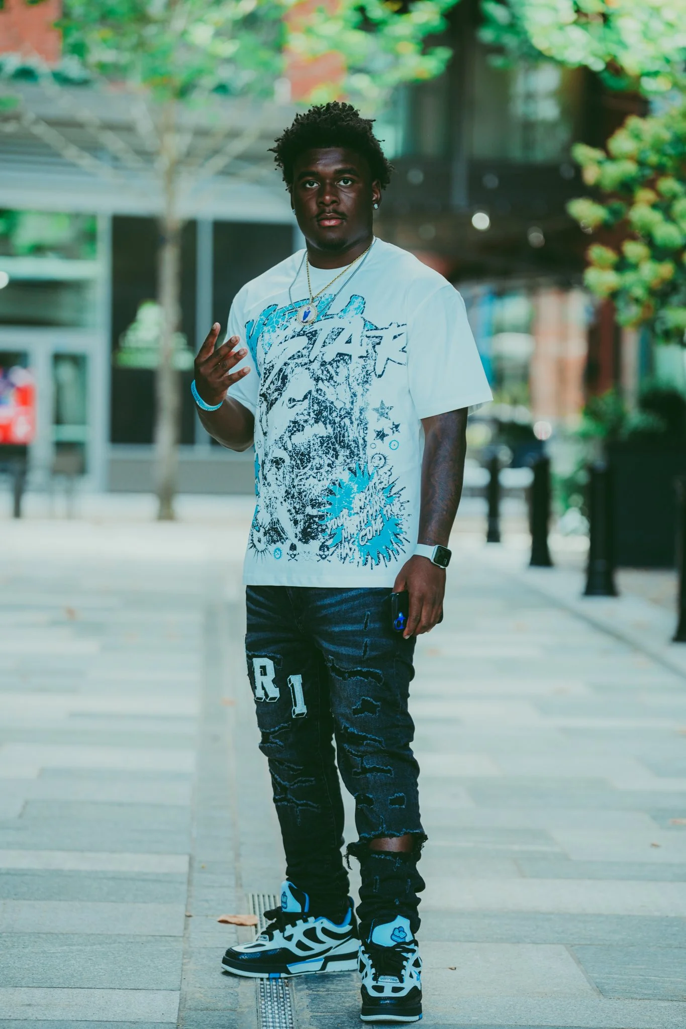 Young man standing on a city sidewalk with trees and buildings in the background, wearing a graphic white t-shirt, ripped jeans, and sneakers, holding a phone in his right hand and making a hand gesture with his left.