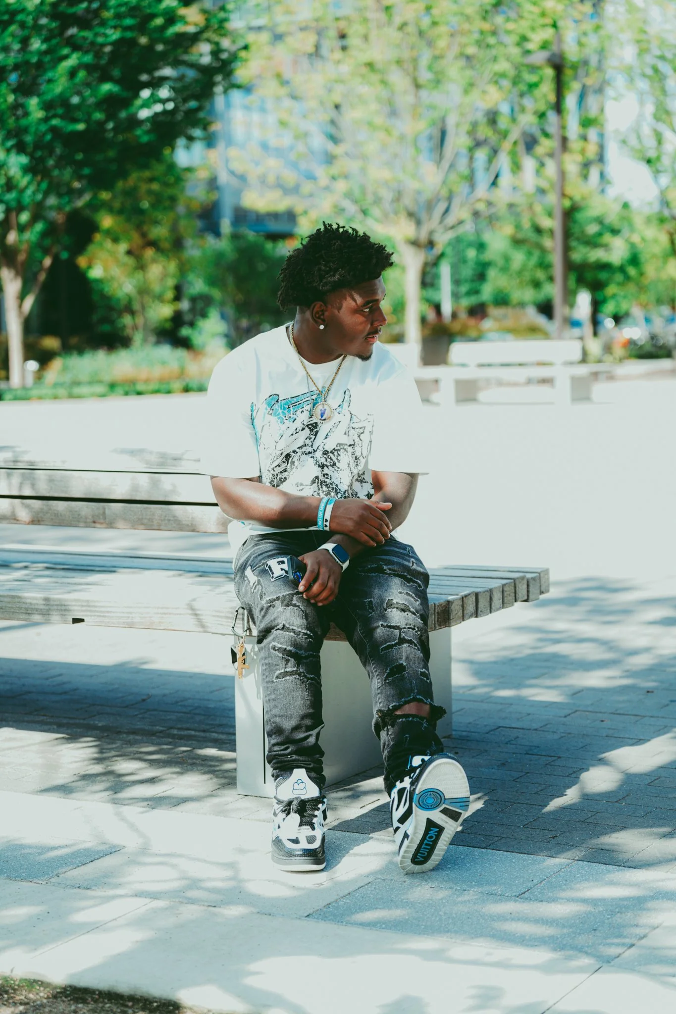 A young man with black, curly hair sitting on a wooden bench outdoors in a park with green trees and clear sky in the background. He is wearing a white graphic T-shirt, distressed black jeans, white sneakers, and accessories including a necklace, ear