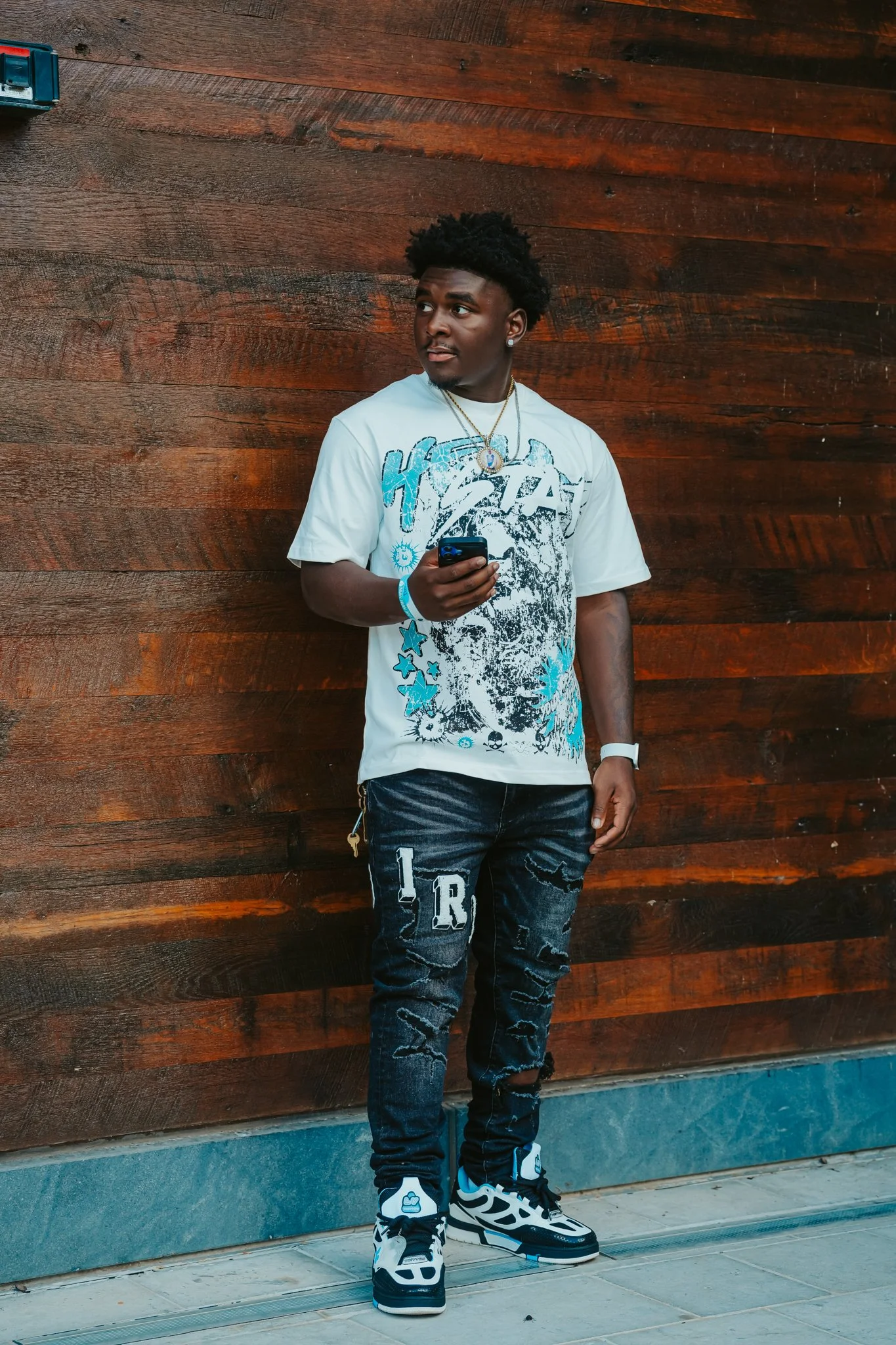 Young man standing against a wooden wall, holding a phone, wearing a white graphic T-shirt, black distressed jeans with patches, and black and white sneakers.