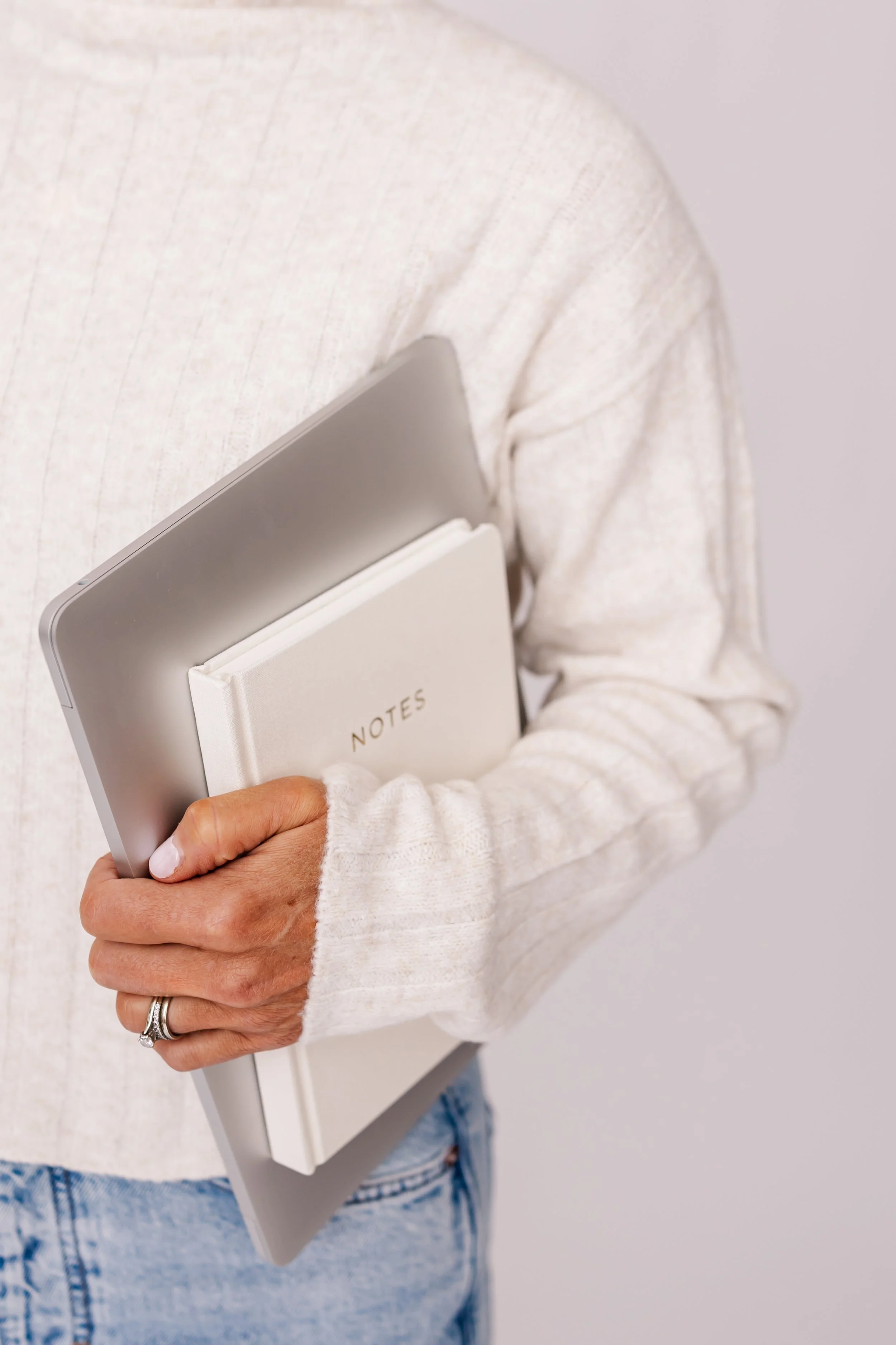 Person wearing a cream-colored sweater holding a tablet, a notebook labeled 'Notes,' and a smartphone, with their left hand showing a wedding ring.