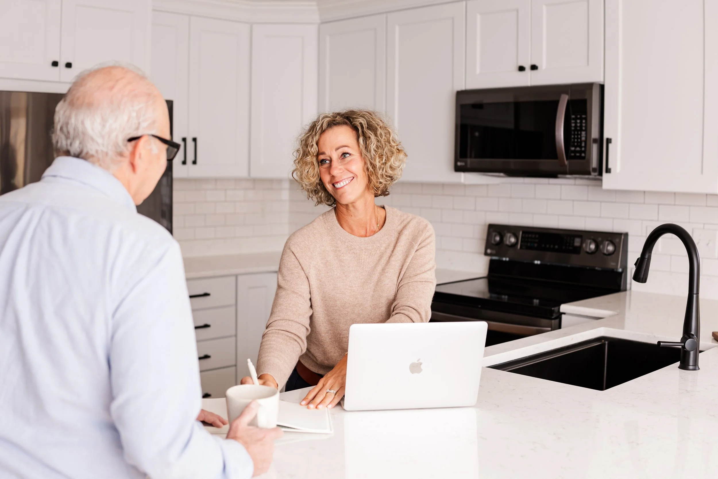 A woman with curly blonde hair smiling while talking to an older man with white hair and glasses in a bright kitchen. The woman is leaning on a white kitchen island with a laptop in front of her, and the man is holding a mug and a notebook.