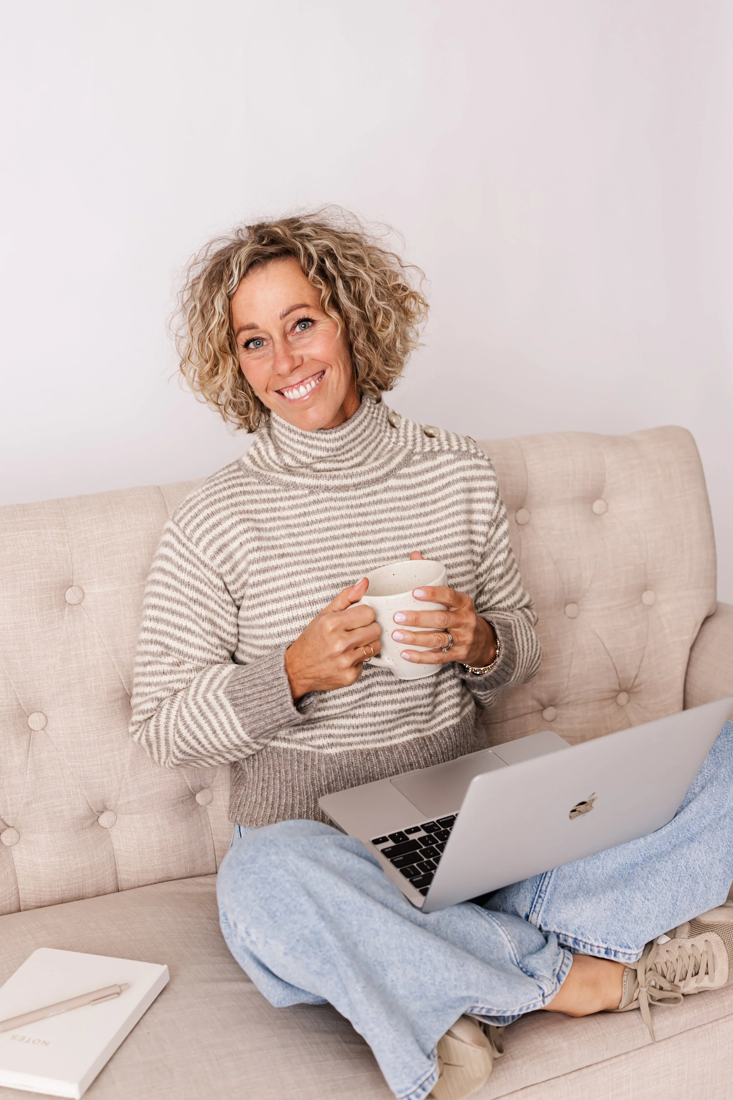 A woman with curly blonde hair sitting cross-legged on a beige sofa, holding a white mug, with a laptop on her lap in a cozy setting.