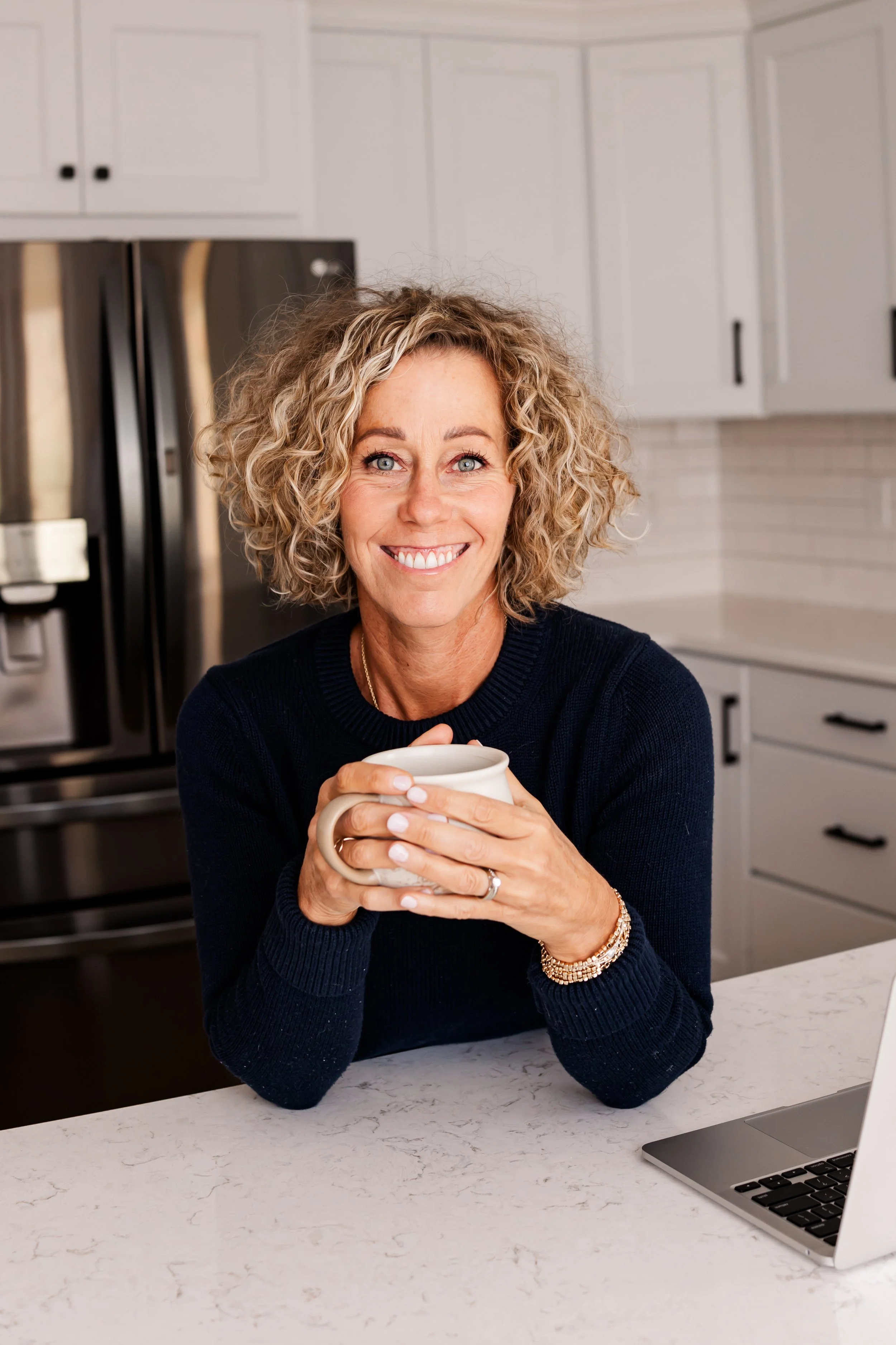 A smiling woman with curly blonde hair holding a mug while sitting at a kitchen counter with a laptop nearby.
