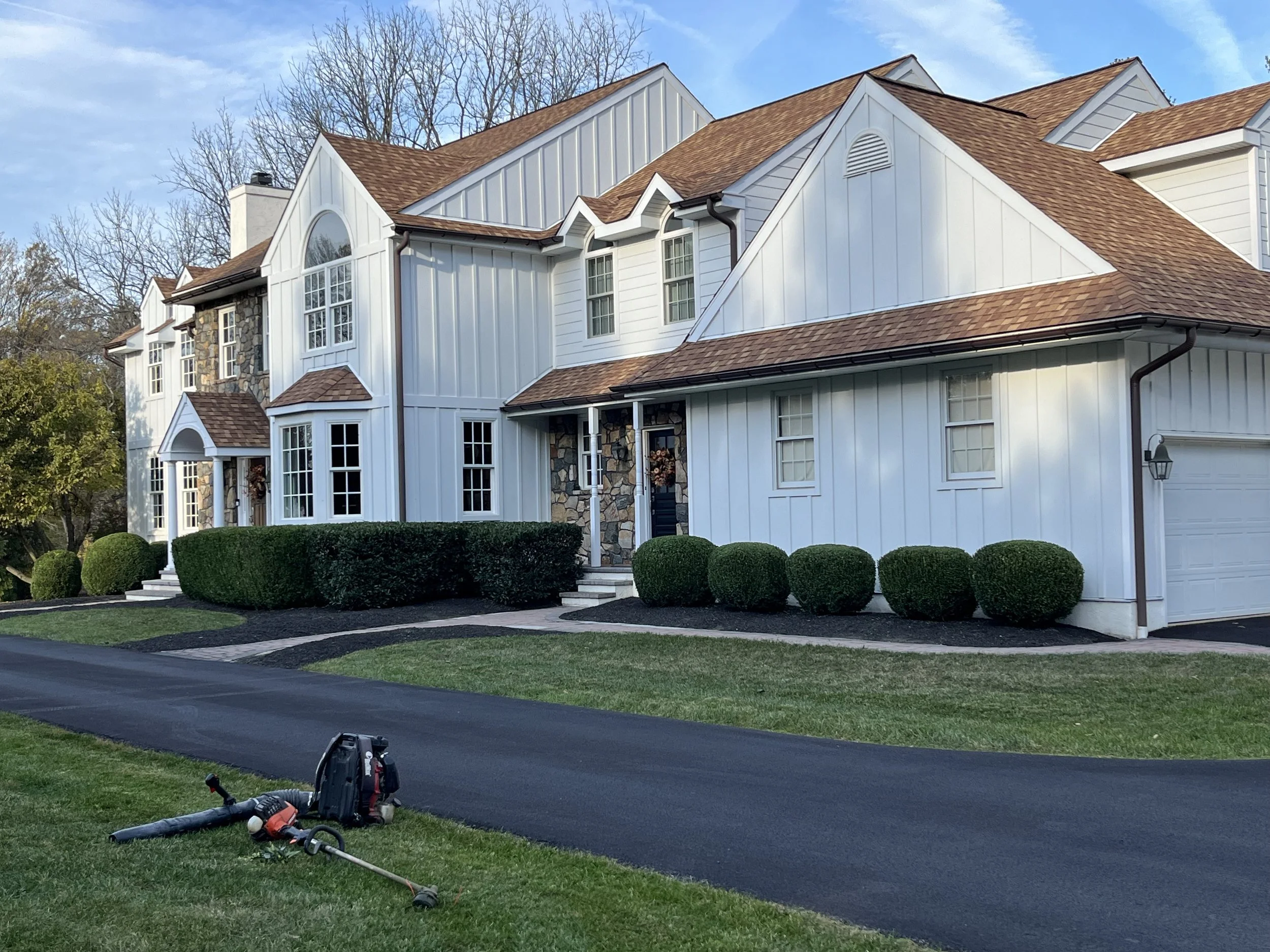 A modern white house with tan shingle roof and stone accents, surrounded by landscaped bushes and a freshly paved driveway, with gardening tools resting on the grass in the front yard.