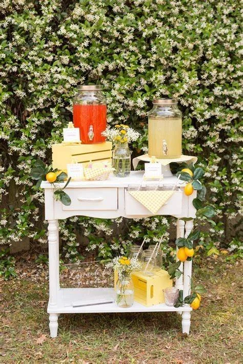 Outdoor refreshment stand with lemonade and iced tea jars, lemons, and yellow floral arrangement on a white table against a lush green shrub background.
