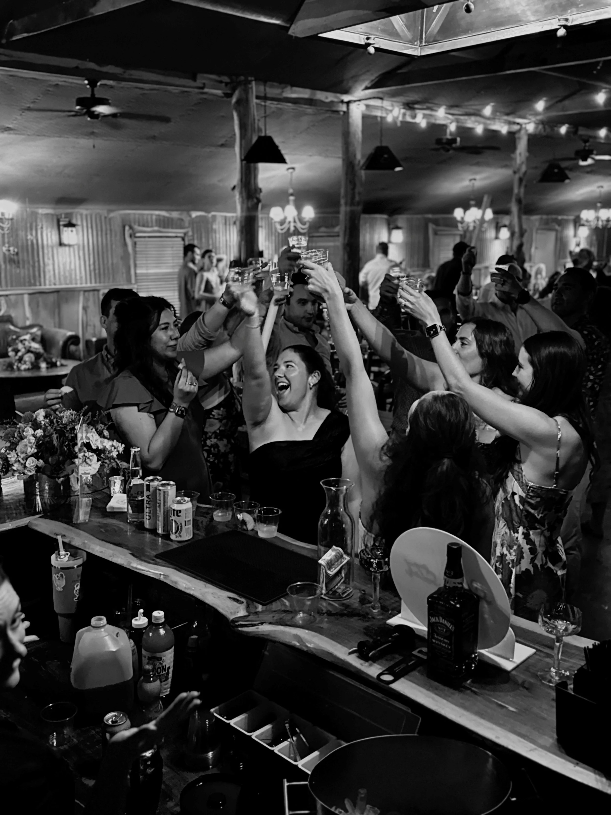 Group of people raising shot glasses in a toast at a lively party or celebration in a rustic bar or restaurant.