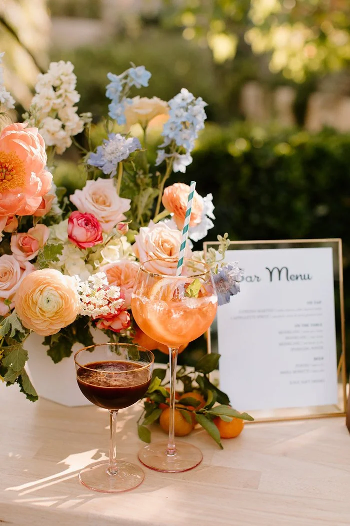 A floral arrangement with pink, peach, and blue flowers, a menu card, and two glasses of beverages on a table in an outdoor setting.