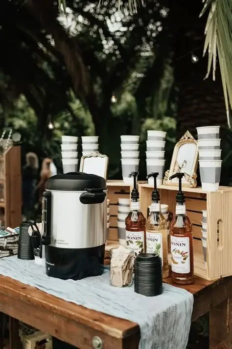 A juice or beverage station with paper cups stacked vertically, bottles of Monin syrup, a large beverage dispenser, and black cups on a wooden table outdoors surrounded by trees.