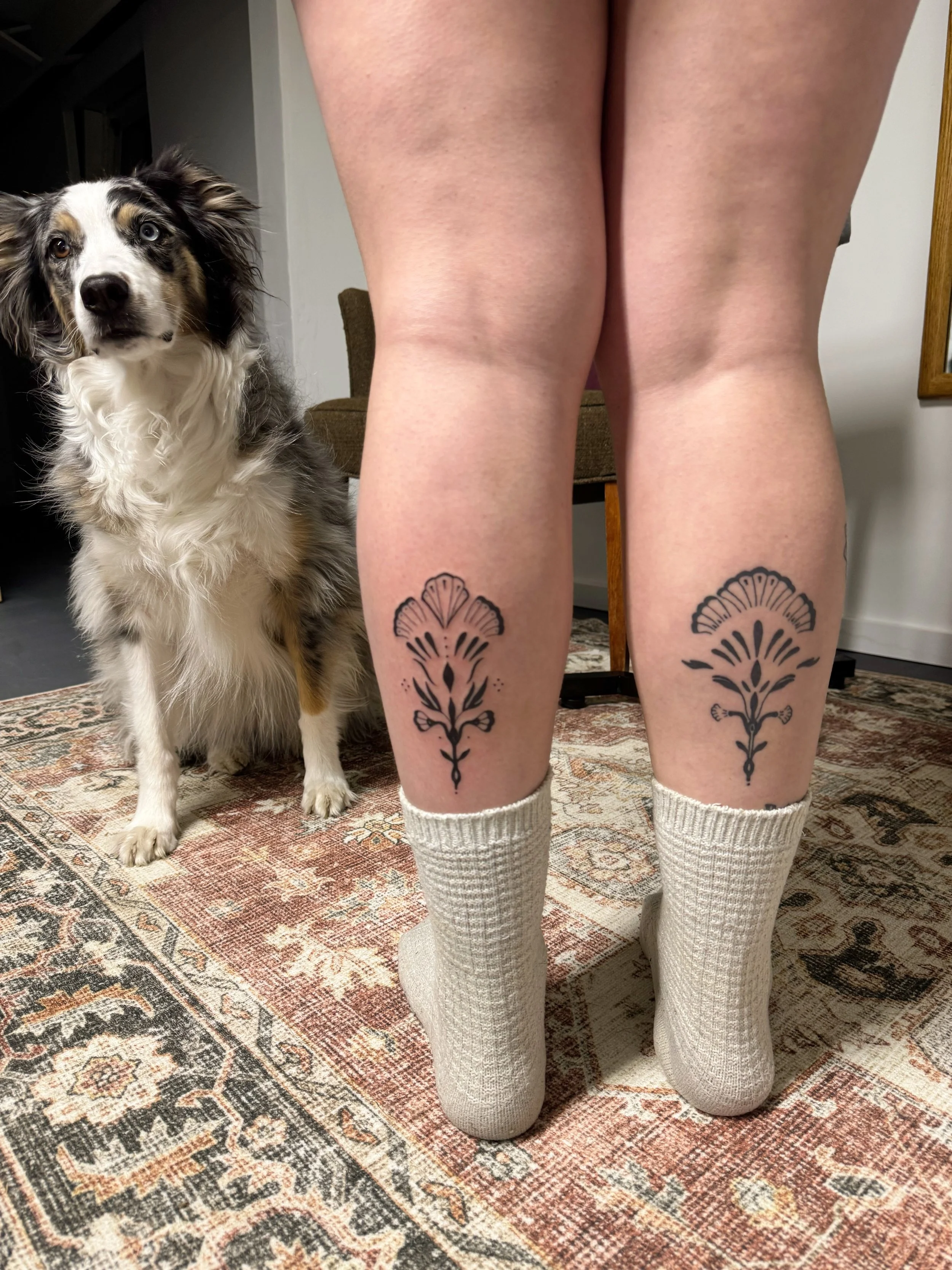 Person standing with their back to the camera, showing their legs with tattoos on the calves. The person is wearing white socks and no shoes. There is an australian shepherd dog with merle coloring, sitting on a patterned rug, looking at the camera.