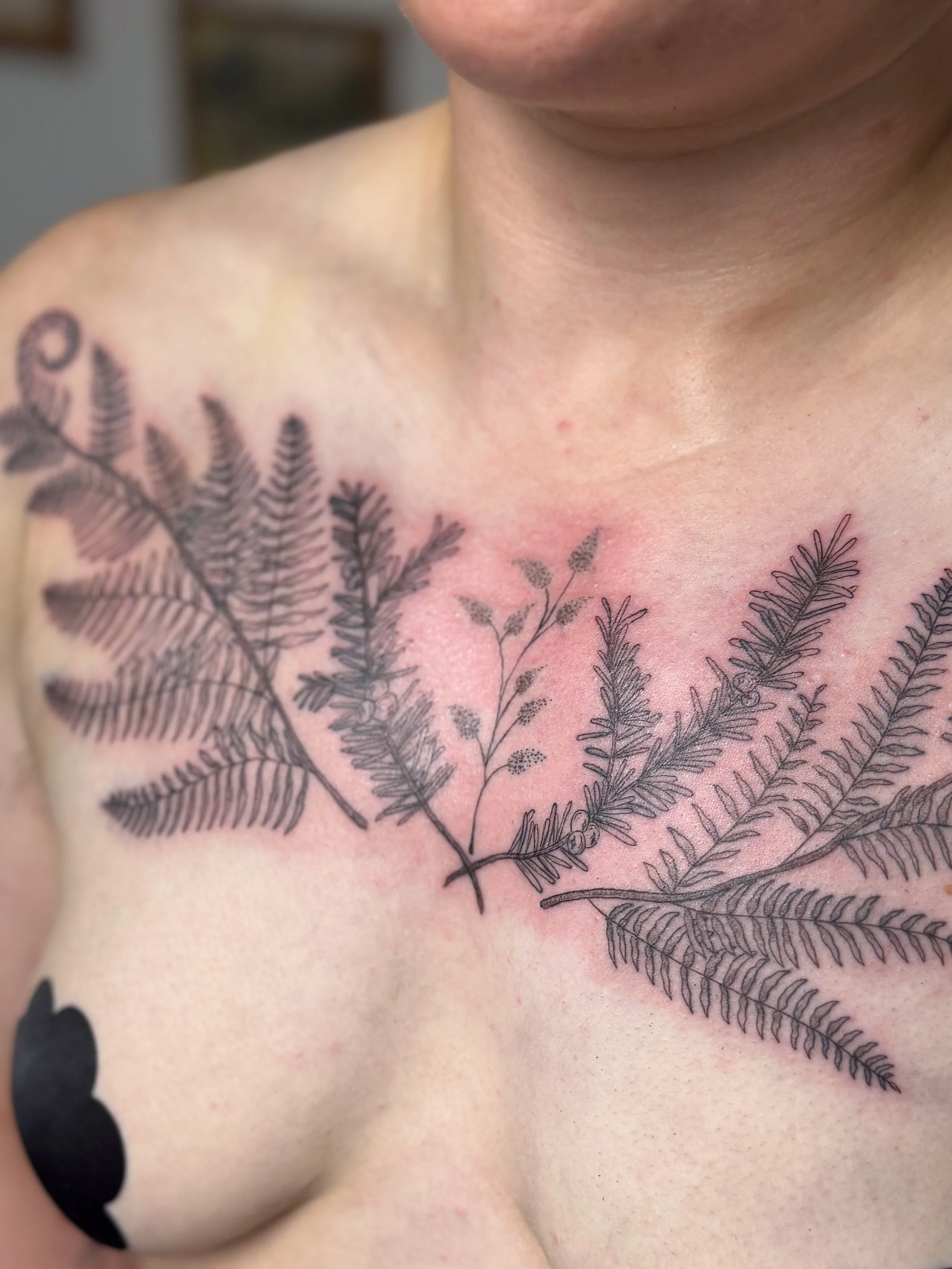 Close-up of a woman's upper chest with a black fern tattoo extending onto her shoulder, and a small black flower on her breast.