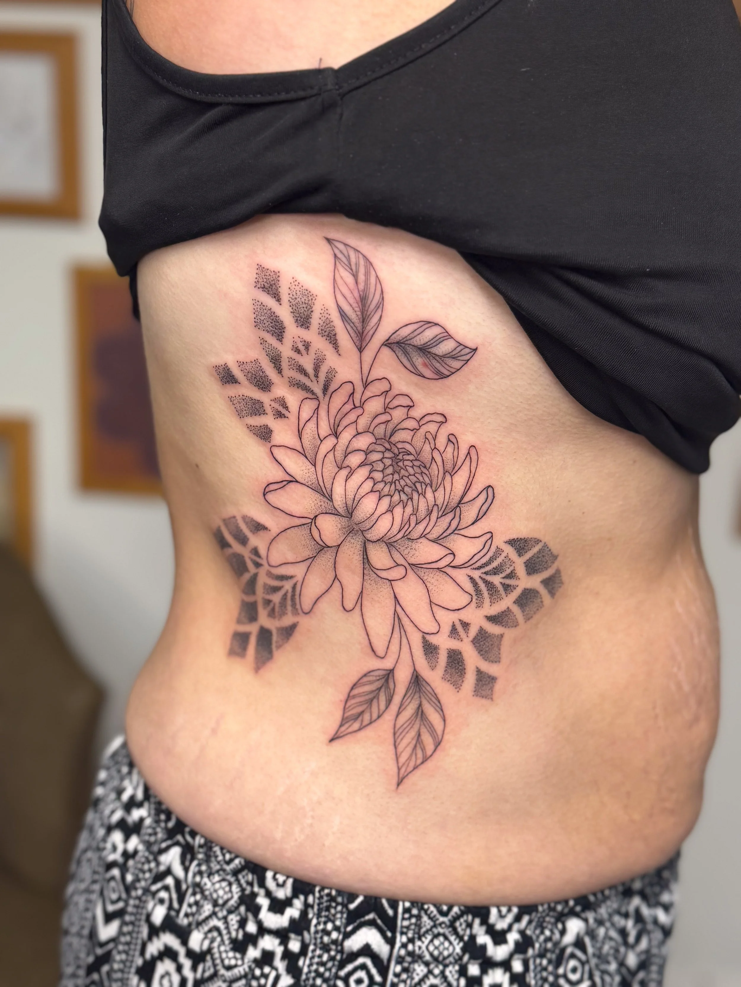 Black line tattoo of a flower with leaves on a person's side torso.