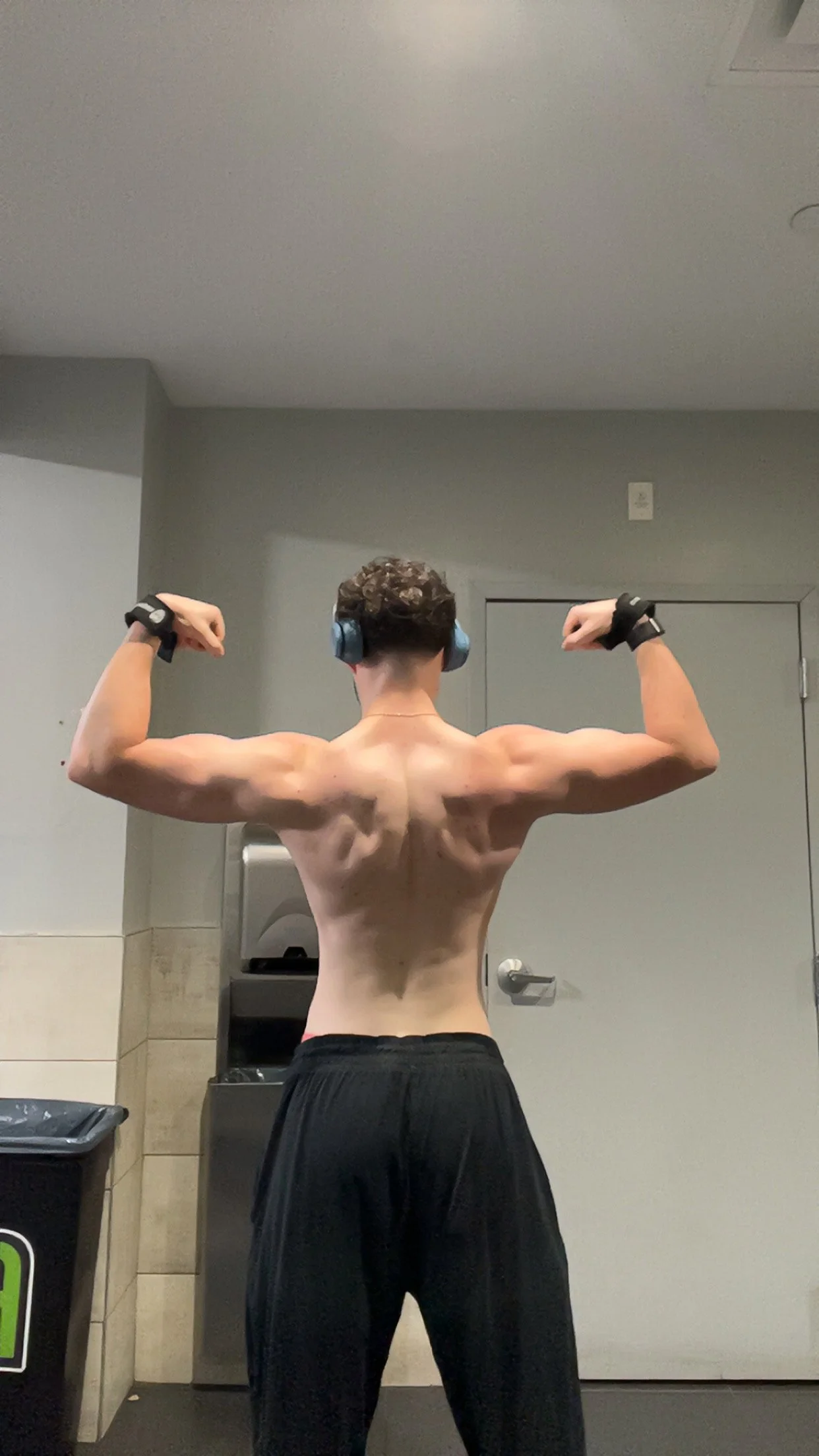 Back view of a shirtless young man flexing his arm muscles at a gym, wearing headphones and black shorts.