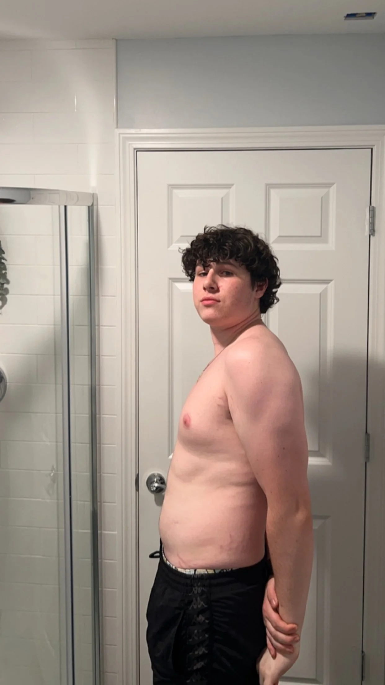 A shirtless young man with curly dark hair standing in a bathroom, facing sideways, with his arms behind his back.