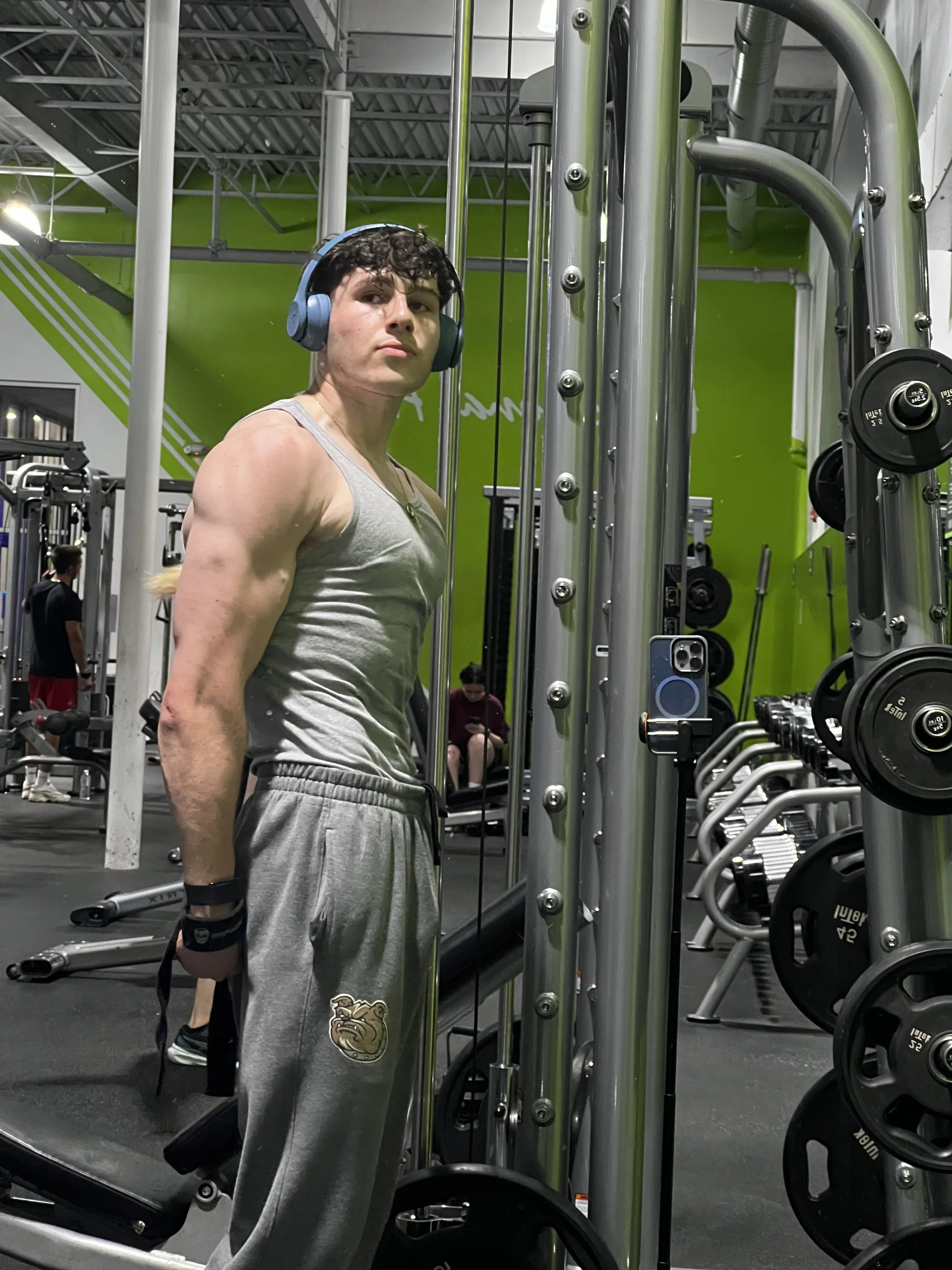 A young man with curly hair wearing a gray tank top, gray sweatpants with a bulldog logo, and headphones, standing in a gym in front of a mirror with weightlifting equipment.