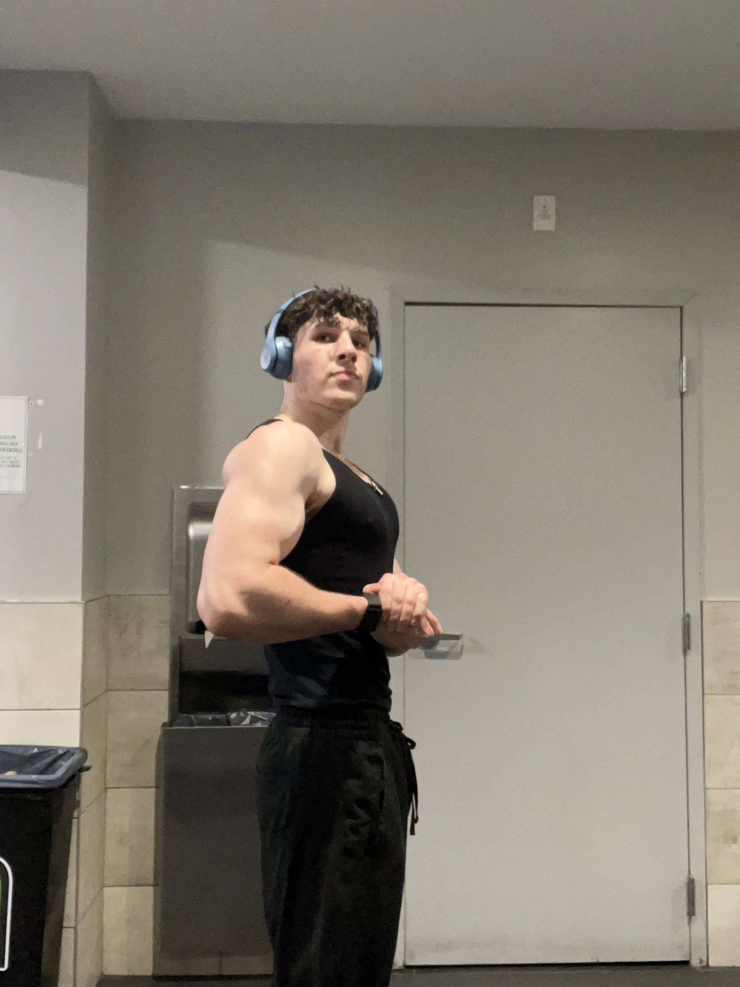 A young man with curly dark hair, wearing a black tank top and black sweatpants, is standing in a gym or locker room. He is wearing blue headphones and has a smartwatch on his left wrist, flexing his right arm to show his muscular bicep.