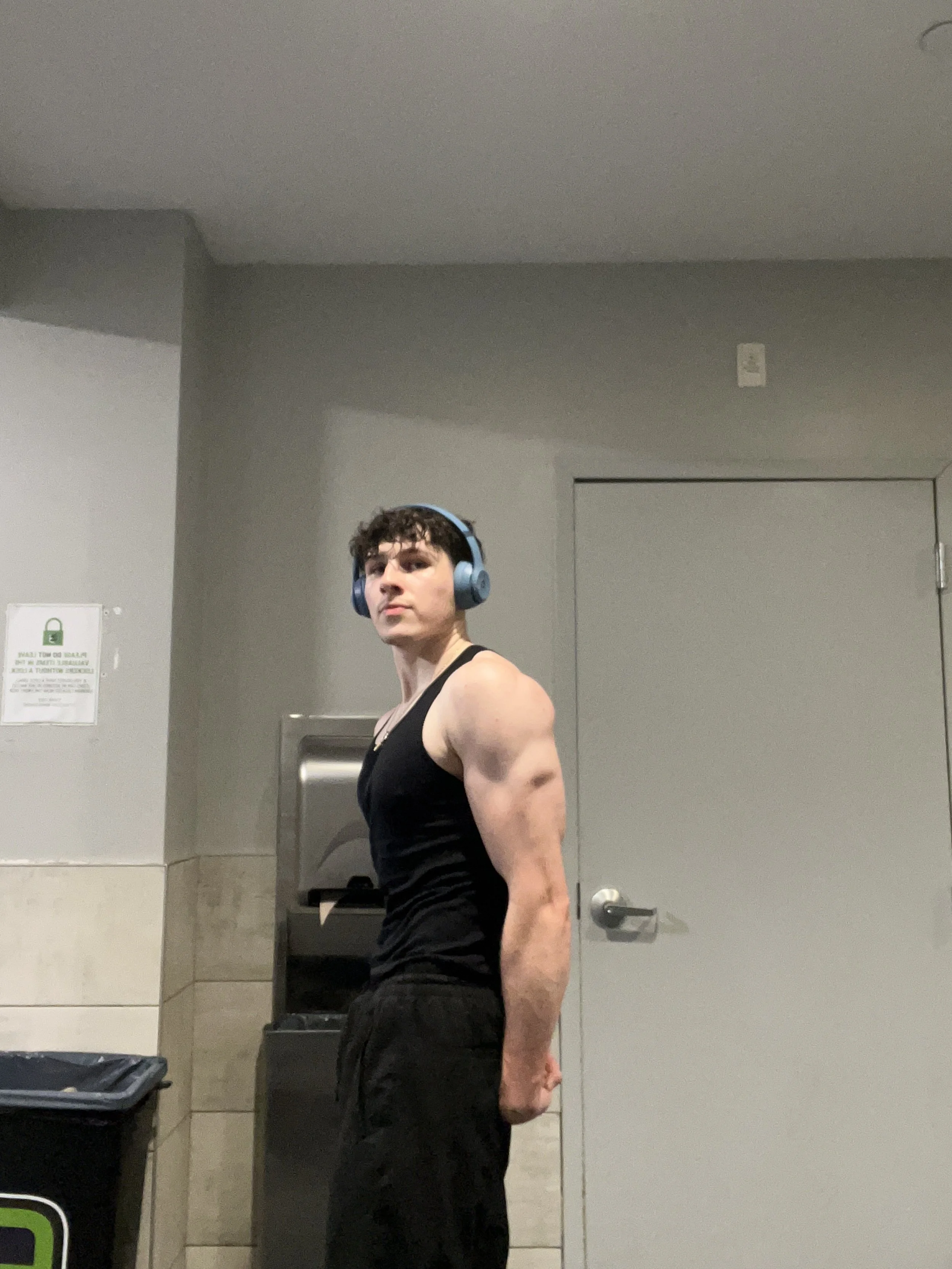 A young man with curly dark hair wearing a black sleeveless shirt, black pants, and blue headphones stands in front of a closed door in a restroom, with a wall-mounted paper towel dispenser behind him.