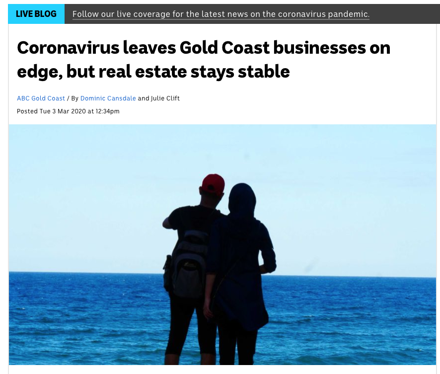 Two people standing on a beach, facing the ocean, one with a red cap and backpack, the other in a dark hoodie and backpack, silhouetted against the blue sea and sky.