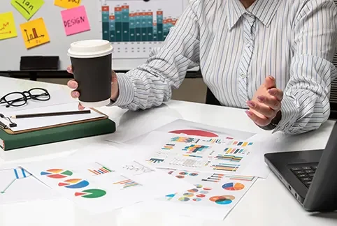 Person sitting at a desk holding a paper cup, with documents, a laptop, and notes on the table, in an office setting.