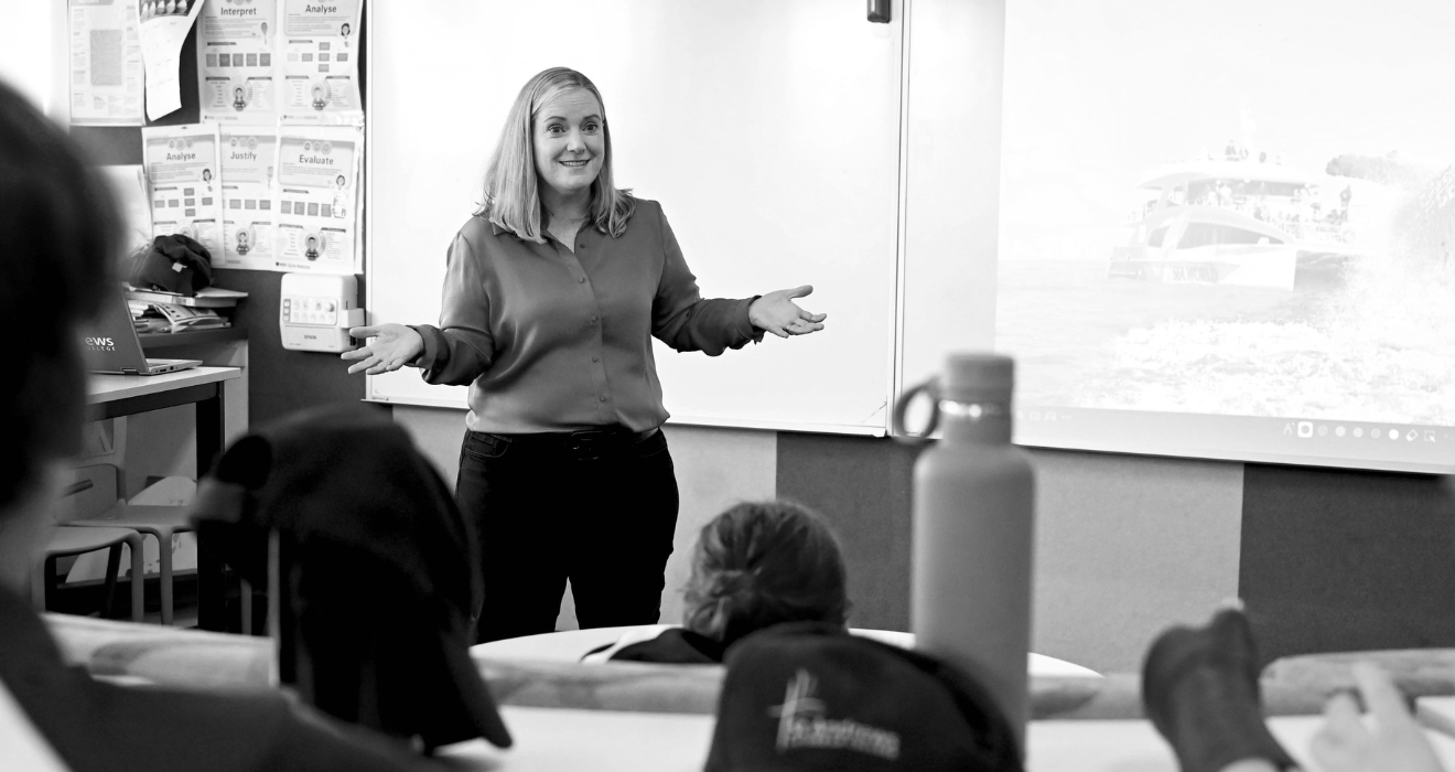 A woman is giving a presentation or lesson in a classroom. She has shoulder-length hair and is wearing a long-sleeve blouse. Students are seated and listening, with some of their heads visible in the foreground. A whiteboard and a projector screen displaying a background image of boats are behind her.
