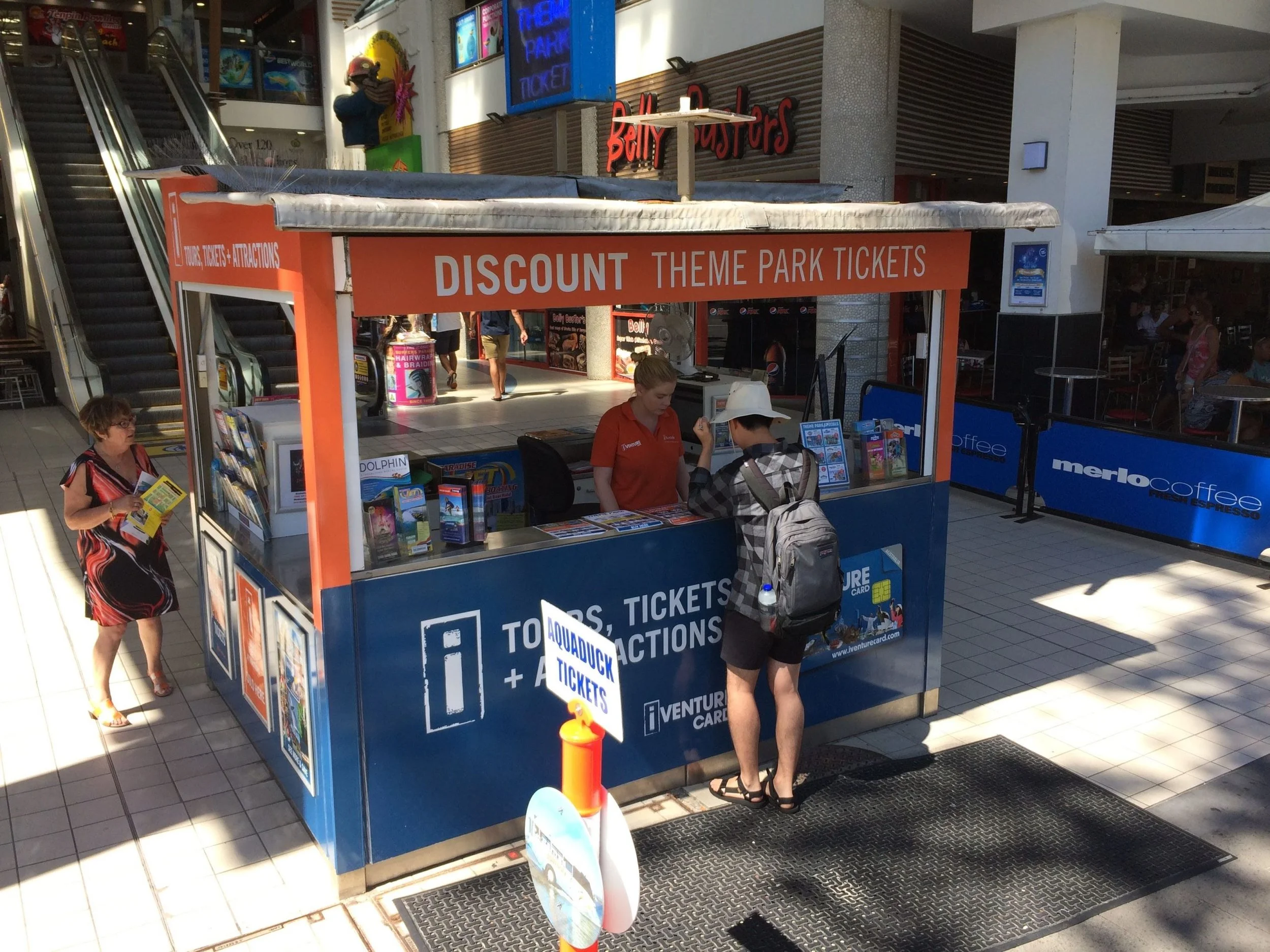 A kiosk in a mall selling tickets for a theme park, with a person at the counter interacting with a worker, a woman holding brochures nearby, and a sign that reads 'AQUADUCK TICKETS' outside the kiosk.