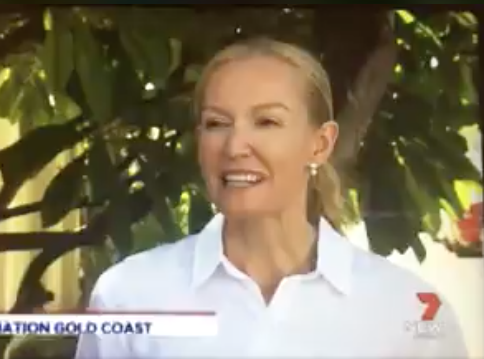 News anchorwoman with blonde hair in a white shirt, speaking outdoors with green leaves in the background.