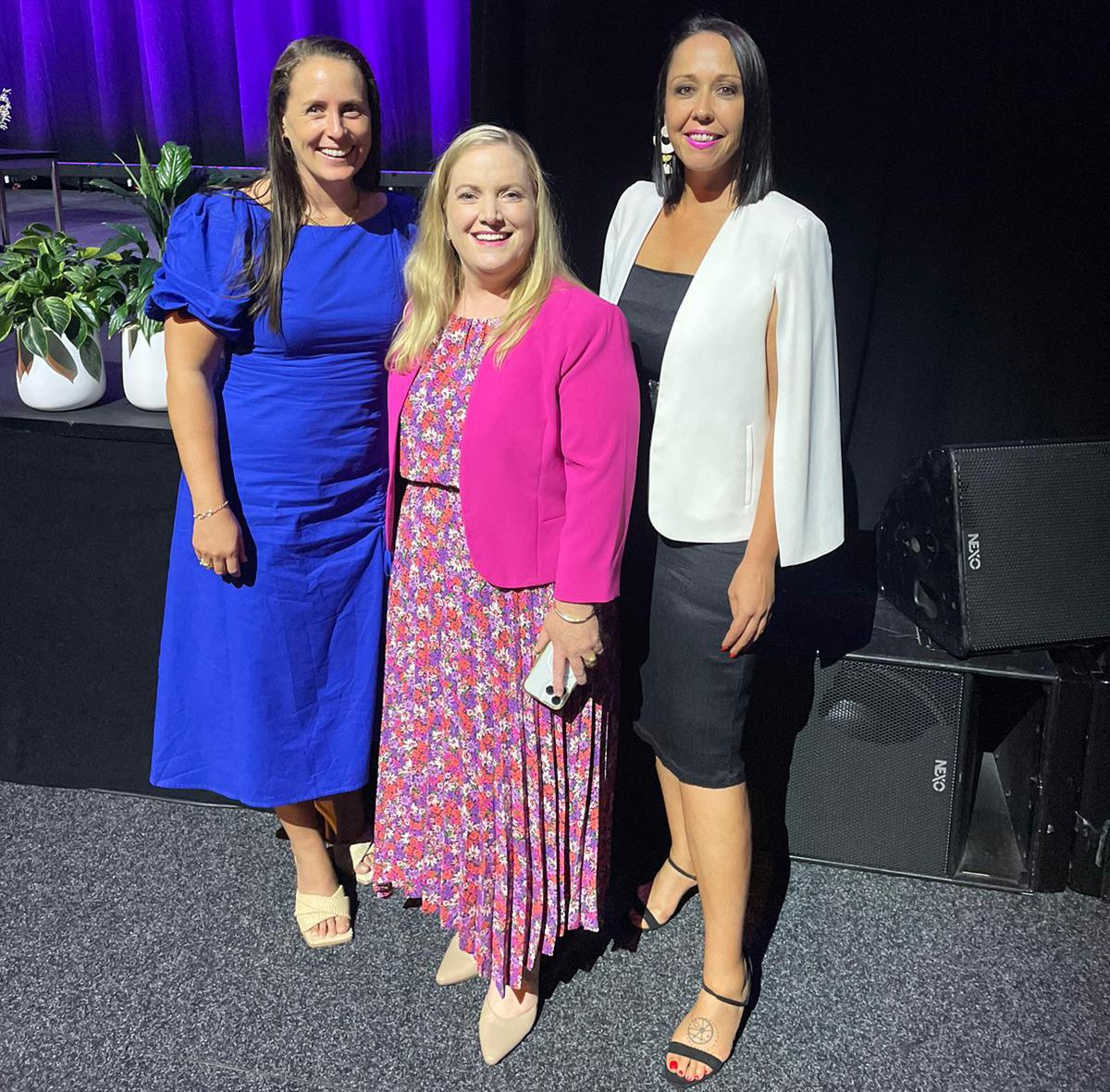 Three women standing together on a stage, dressed in formal attire, smiling for the photo. They are in front of a black backdrop with purple lighting and potted plants.