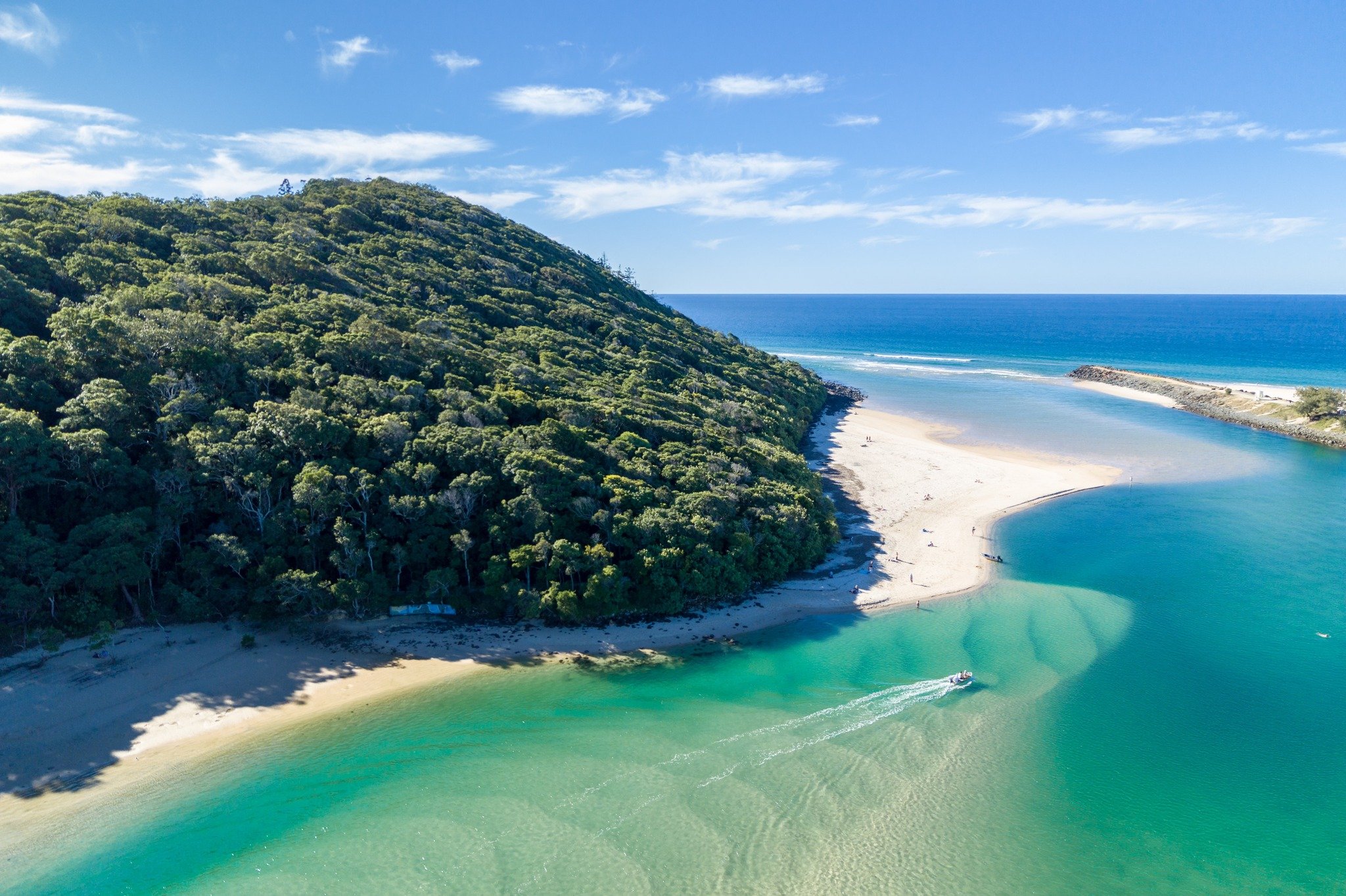 Aerial view of a coastal area with a green forested hill on the left, a sandy beach, and a calm turquoise river flowing into the ocean under a partly cloudy sky.