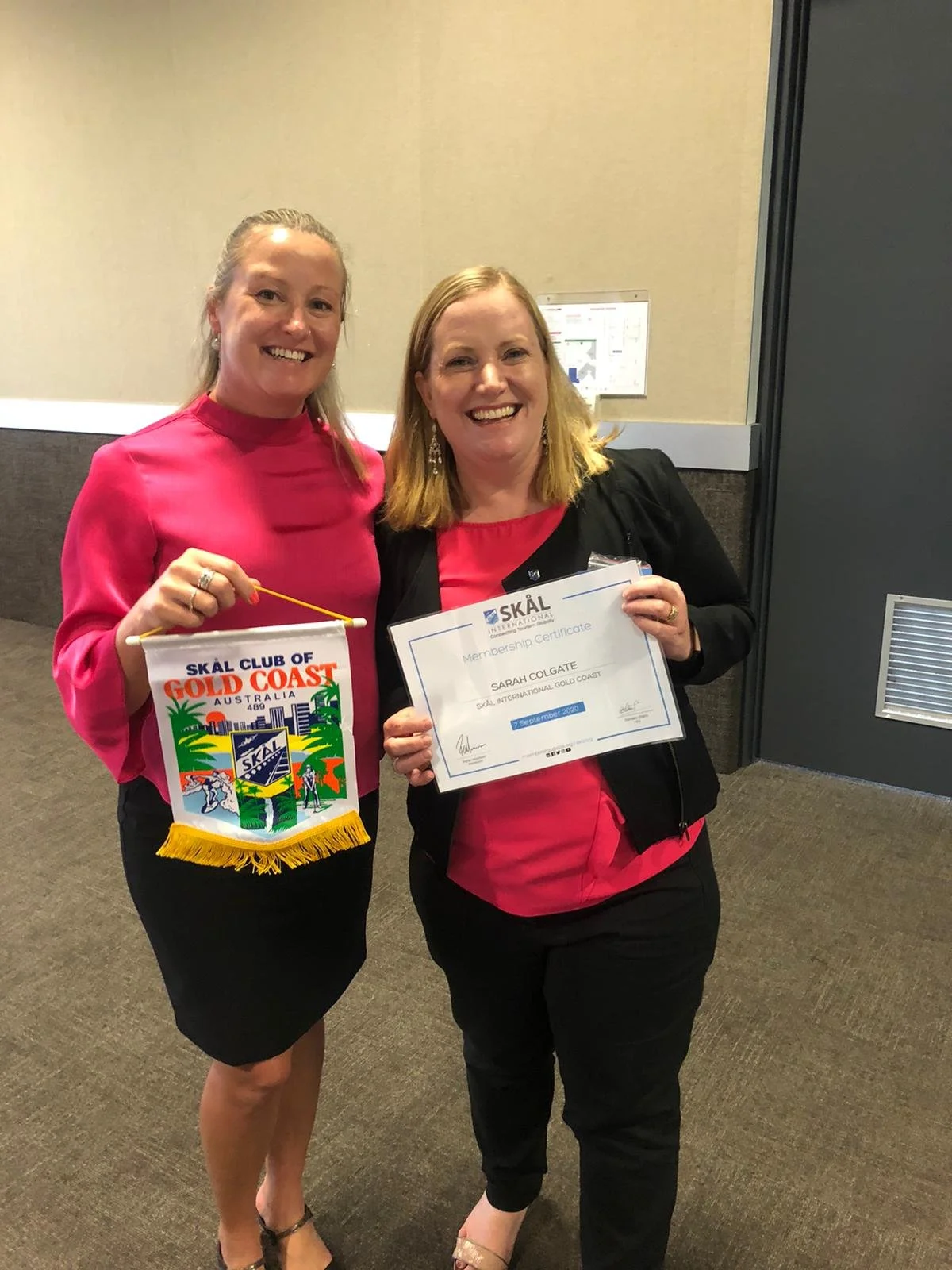 Two women smiling indoors, one holding a colorful banner and the other holding a certificate.