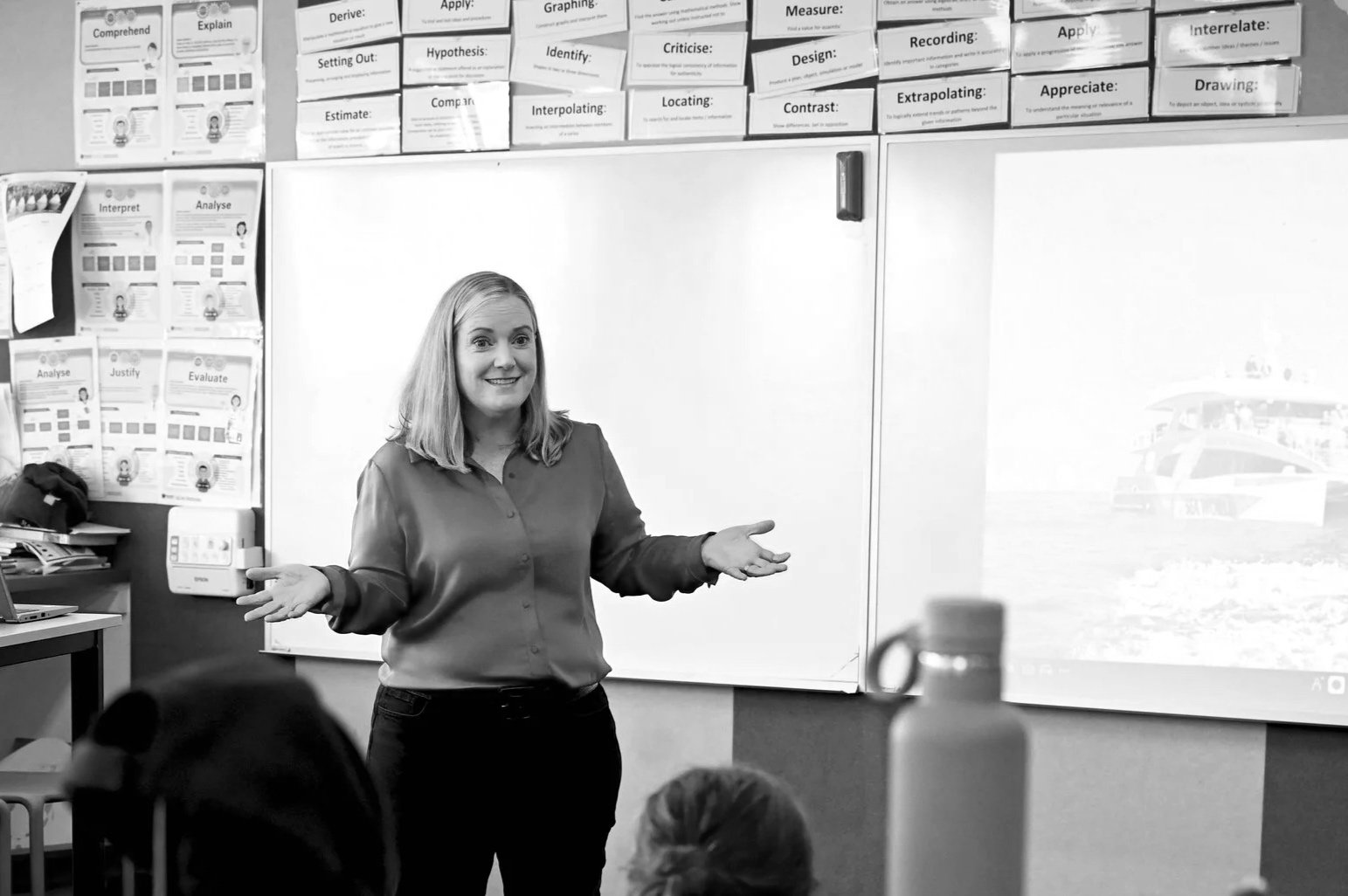 A woman standing in front of a classroom whiteboard, addressing students, with classroom posters and a water bottle visible in the foreground.