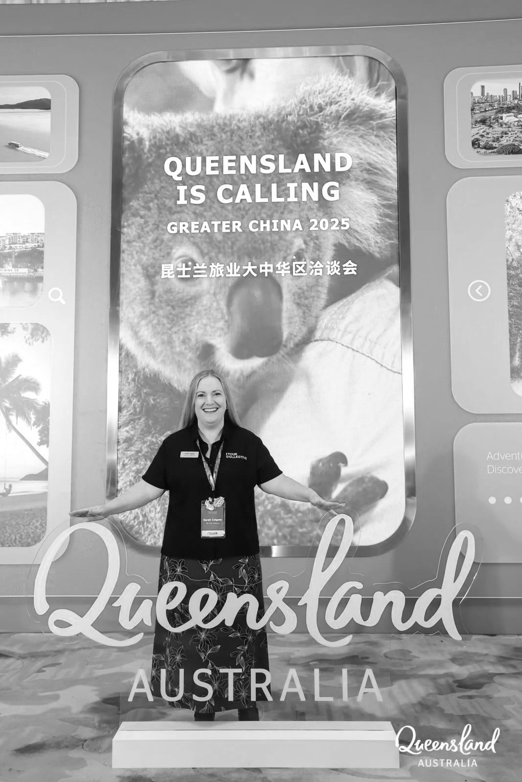A woman standing in front of a large sign that says "Queensland is Calling Greater China 2025" with a picture of a koala bear in the background. The sign also has Chinese characters on it. The woman is smiling and posing with her arms outstretched.