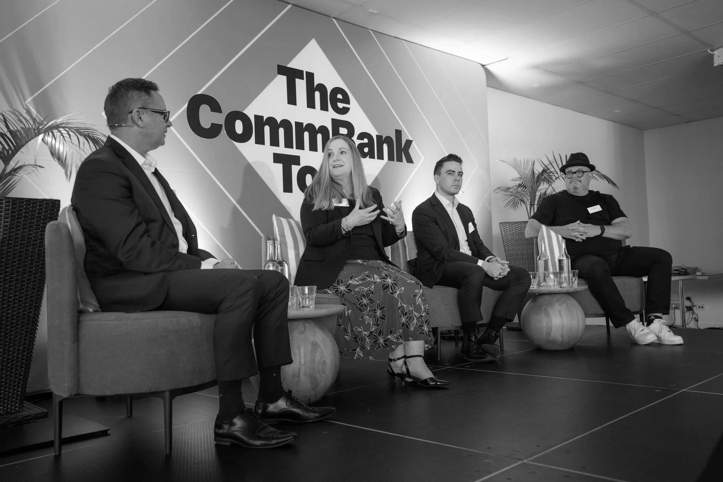 A panel of four people seated on stage during a discussion at The CommBank To event. The woman in the center is speaking, gesturing with her hands. The stage backdrop features the event logo and some potted plants.