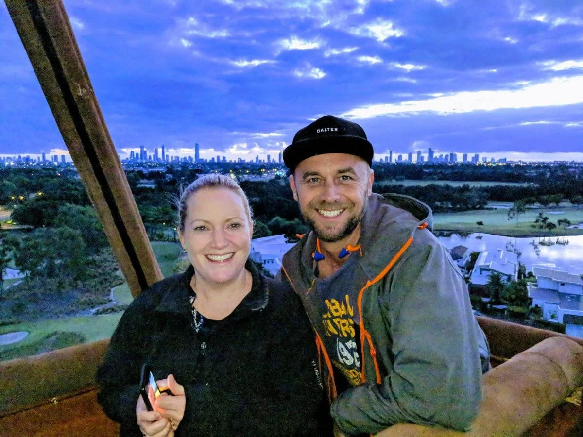 A smiling woman and man are taking a selfie on a hot air balloon ride during sunset, with a city skyline and lush greenery in the background.