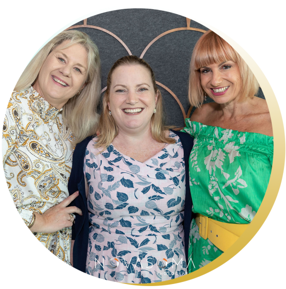 Three women smiling and standing close together in front of a decorative background, dressed in colorful shirts.