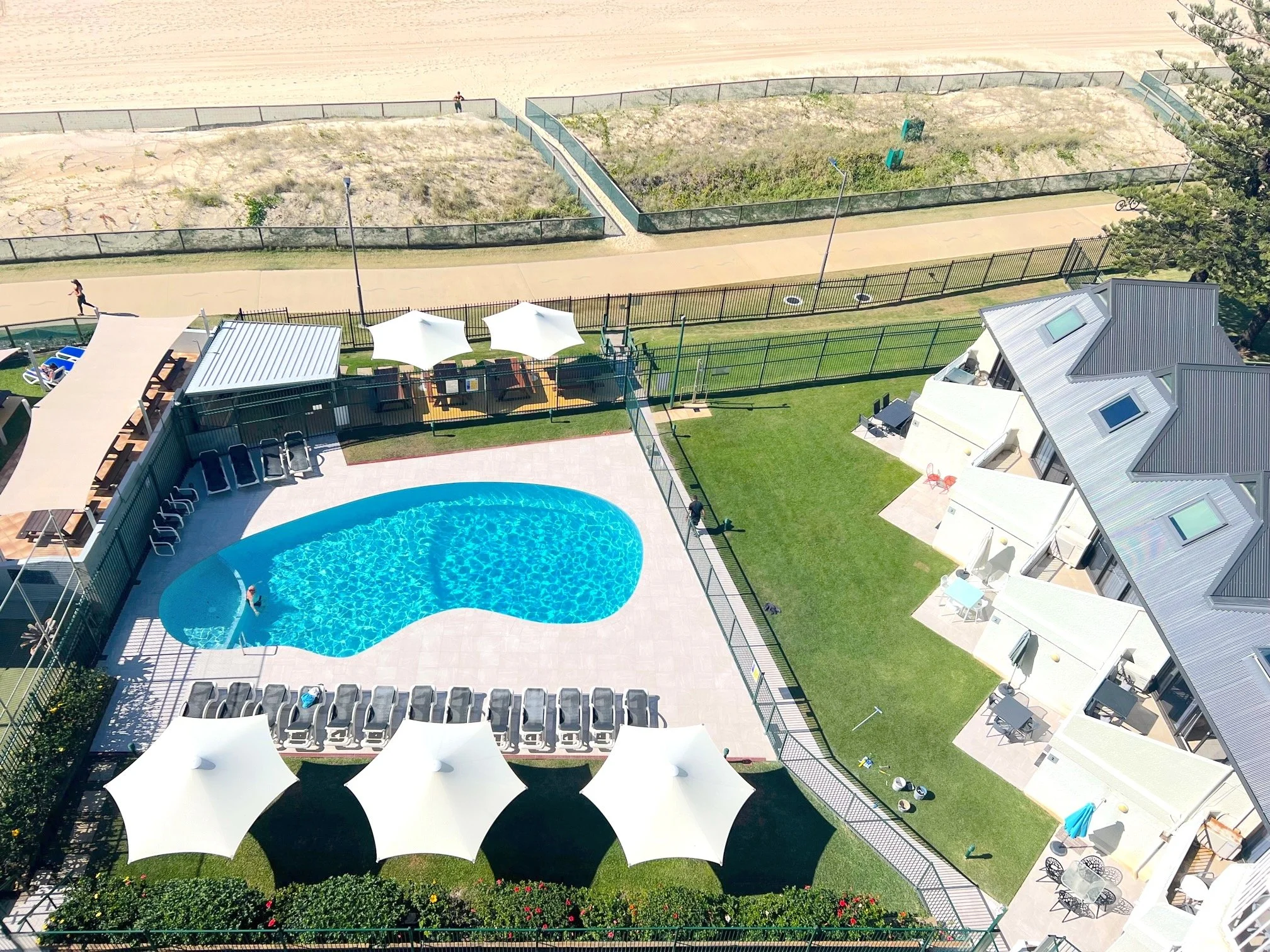 An aerial view of a residential apartment complex with a swimming pool, lounge chairs, umbrellas, and a yard, adjacent to a walking and biking path.