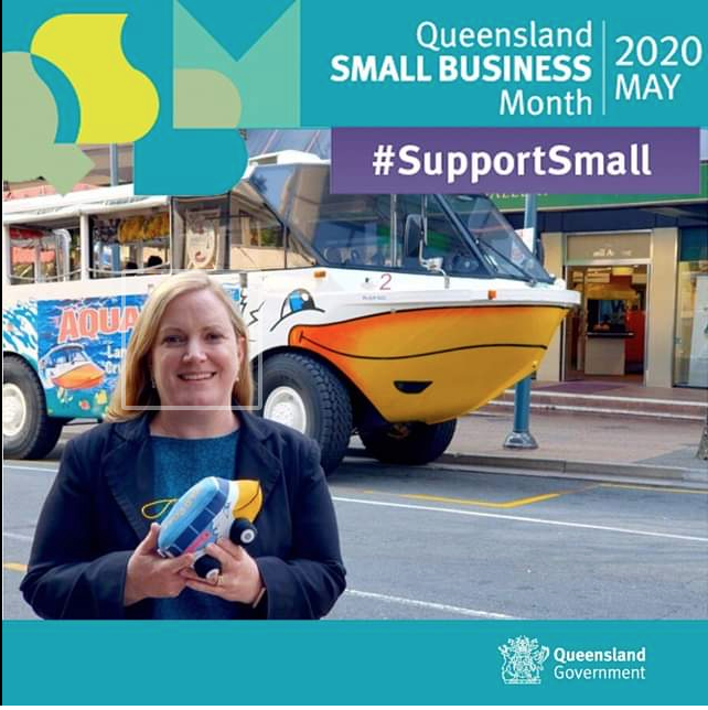 A woman holding a small toy boat in front of a houseboat with a high bow designed to look like a smiling face, in an urban setting, with a message supporting small businesses during Queensland Small Business Month in May 2020.