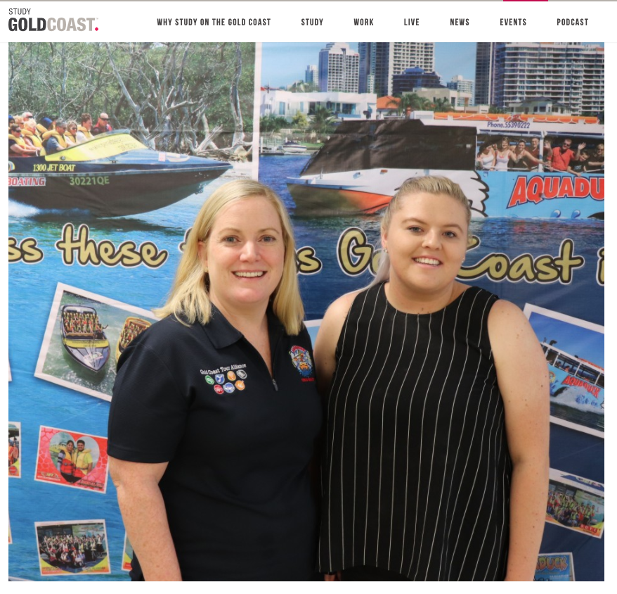Two women standing in front of a promotional backdrop for the Gold Coast, featuring images of boats and a city skyline. The woman on the left has blonde hair, wears a navy polo shirt with pins, and is smiling. The woman on the right has blonde hair,