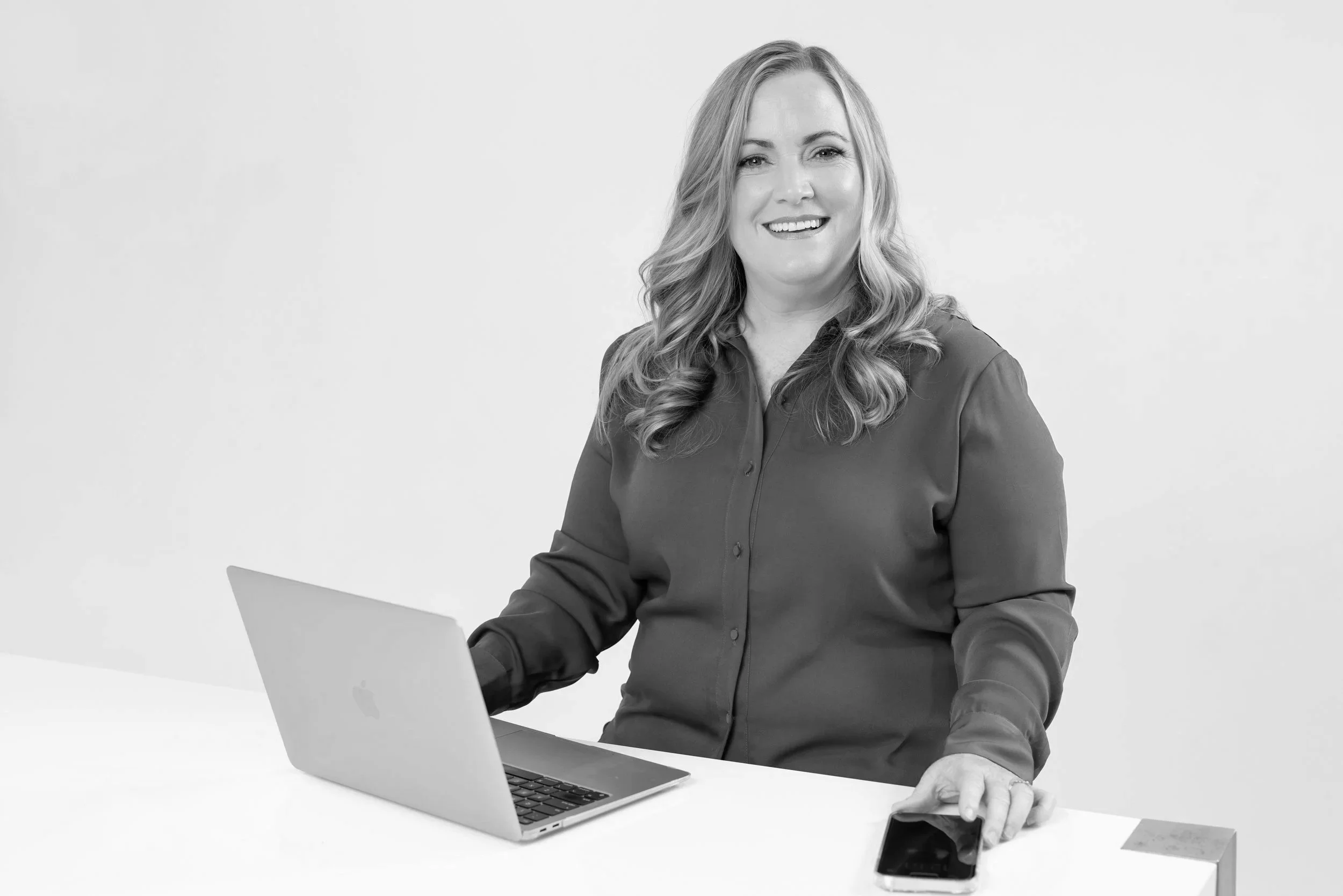 A woman with wavy hair smiling while sitting at a desk with a laptop and a smartphone, in a black-and-white photo.
