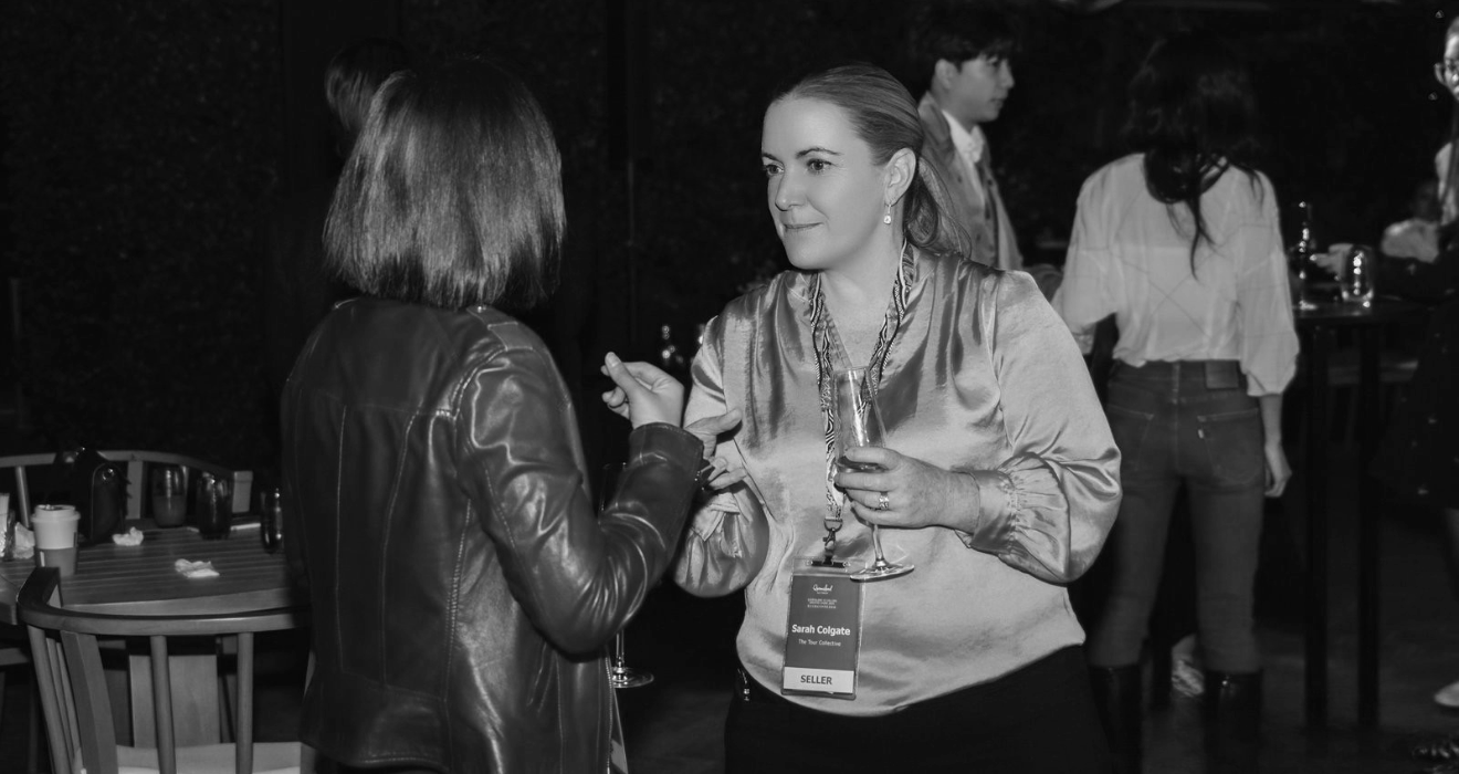 Two women engaging in conversation at a social event, one wearing a leather jacket and the other holding a wine glass with a name tag that reads "Sarah Colgate, The Star Collecting, SELLER". In the background, several other people are visible, some standing around tables.