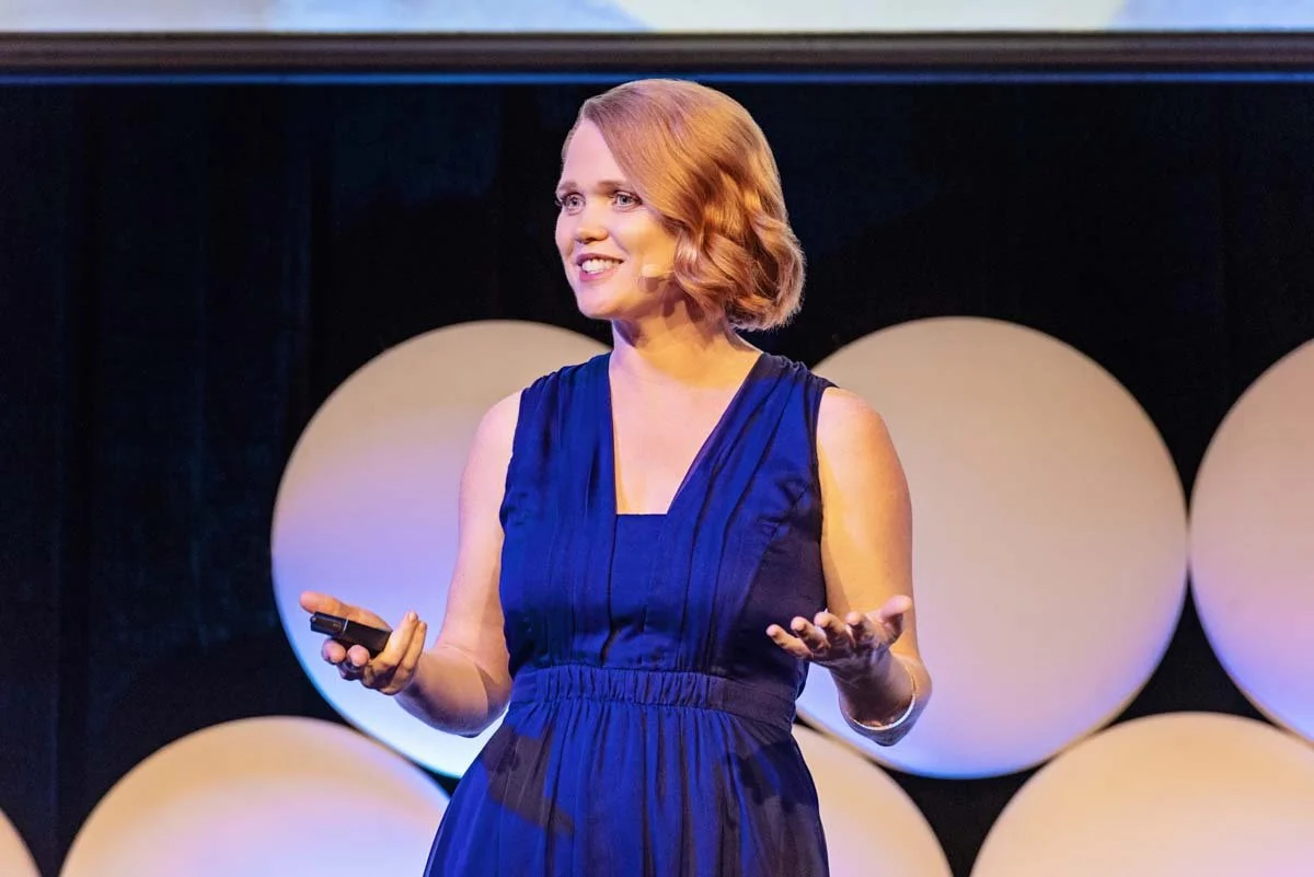 Woman with short red hair wearing a sleeveless dark blue dress, speaking on stage with circular background decor.