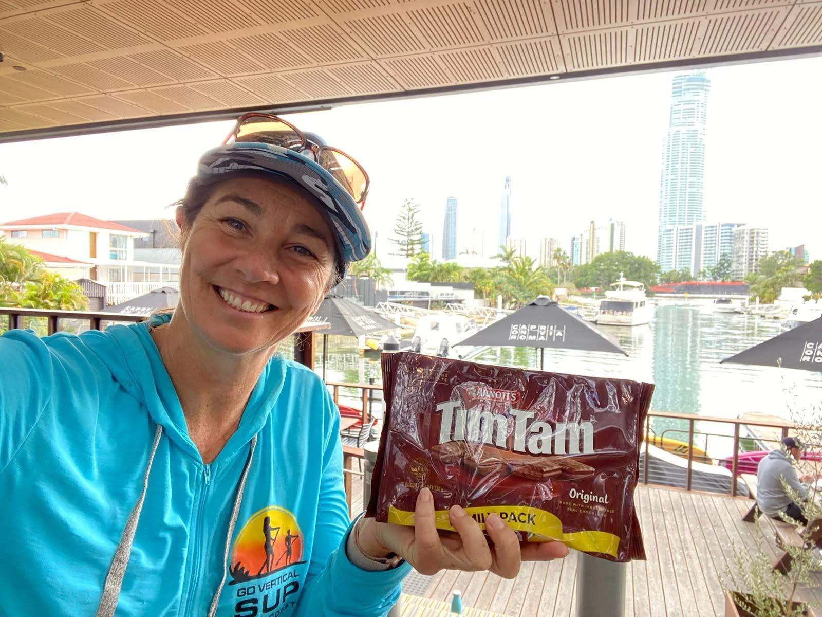 A smiling woman wearing a blue jacket and cap taking a selfie outdoors near a marina with boats, buildings, and trees in the background, holding a package of Tim Tam cookies.