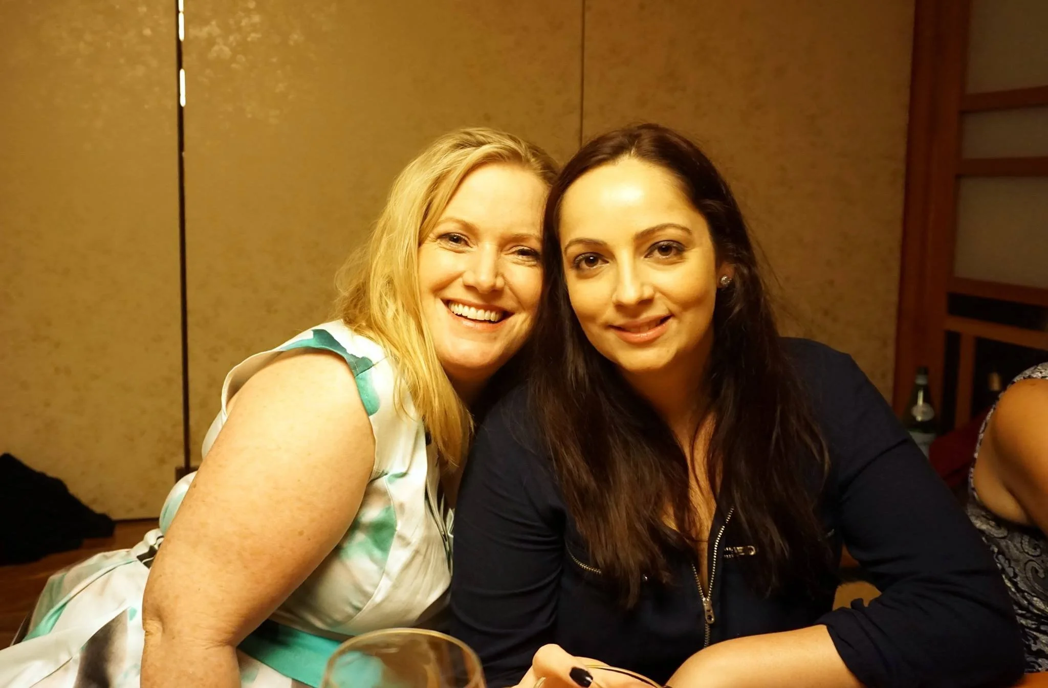 Two women smiling and sitting close together at a table in a warmly lit room.