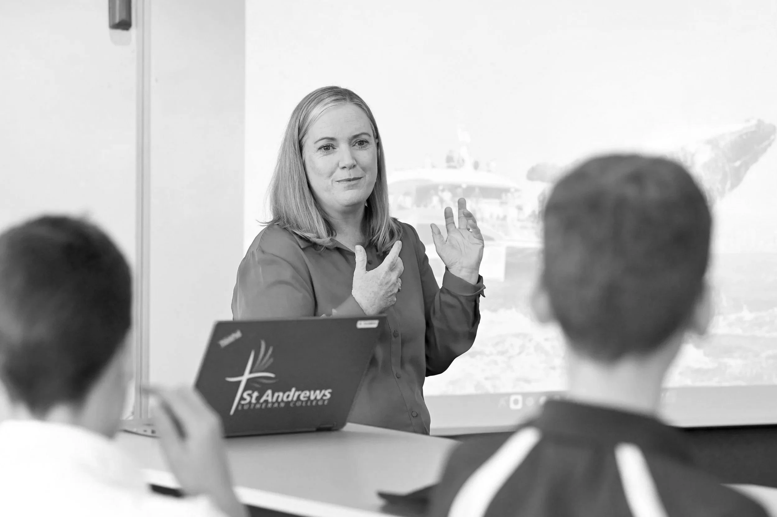 A woman in a long-sleeve shirt giving a presentation to two students in a classroom. The woman is standing behind a laptop with a 'St. Andrews' logo. The students are seated, facing her, with their backs to the camera.