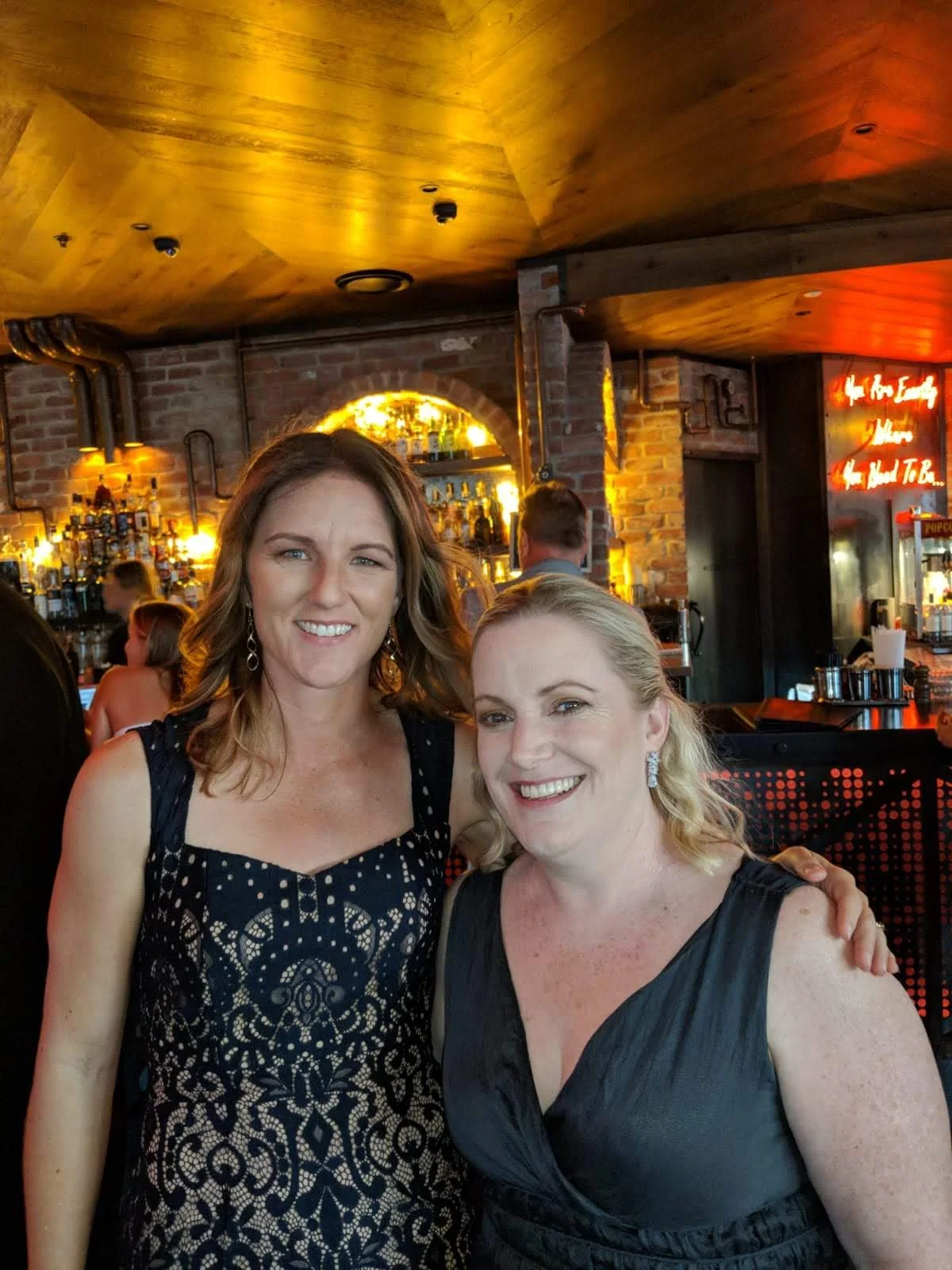 Two women smiling at a bar or restaurant with a warm, brick interior and bottles on shelves in the background.