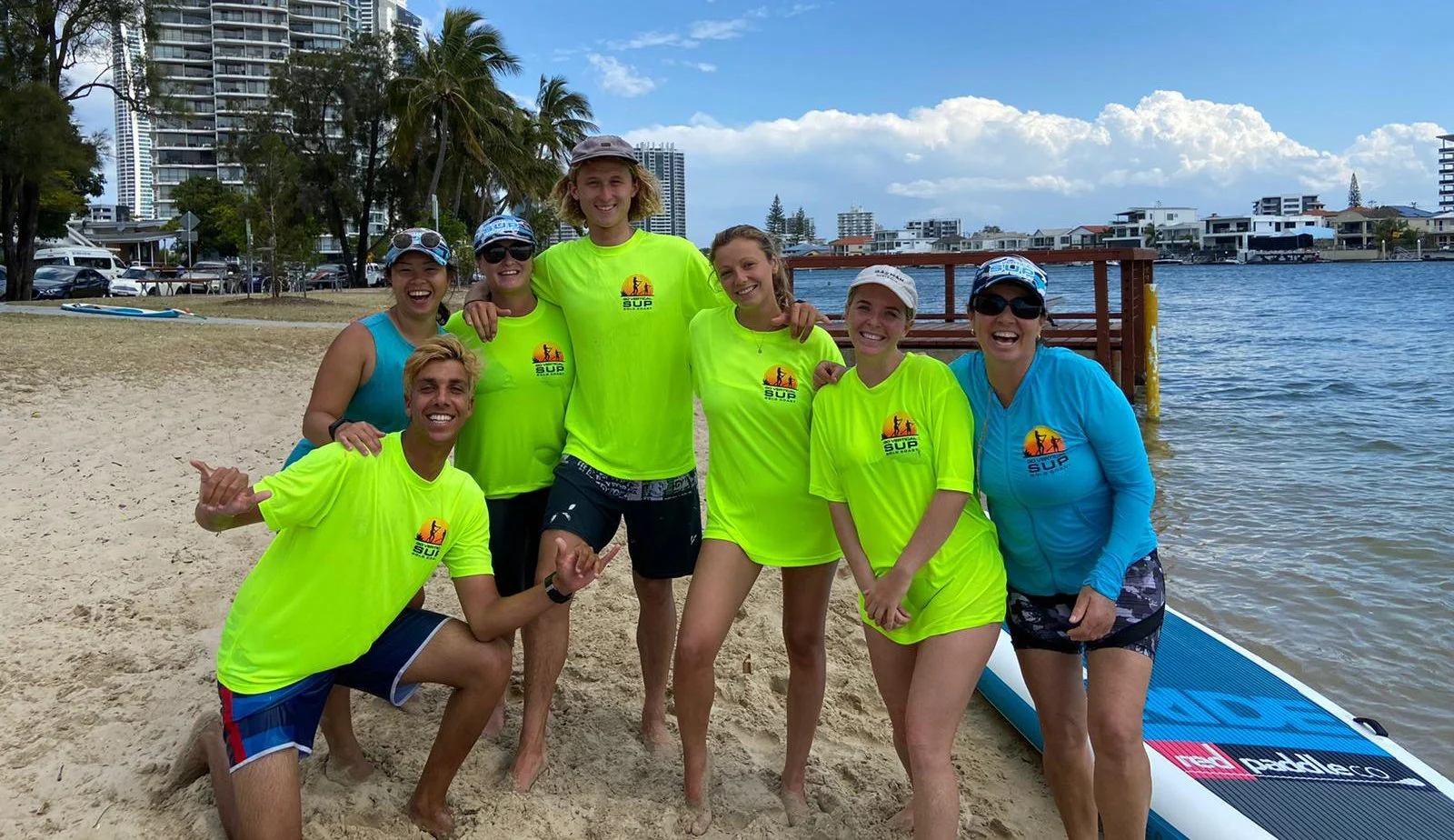 Group of seven people on a sandy beach, wearing matching bright yellow and blue apparel with a SUP logo, smiling and posing in front of a paddleboard by the water with buildings in the background.