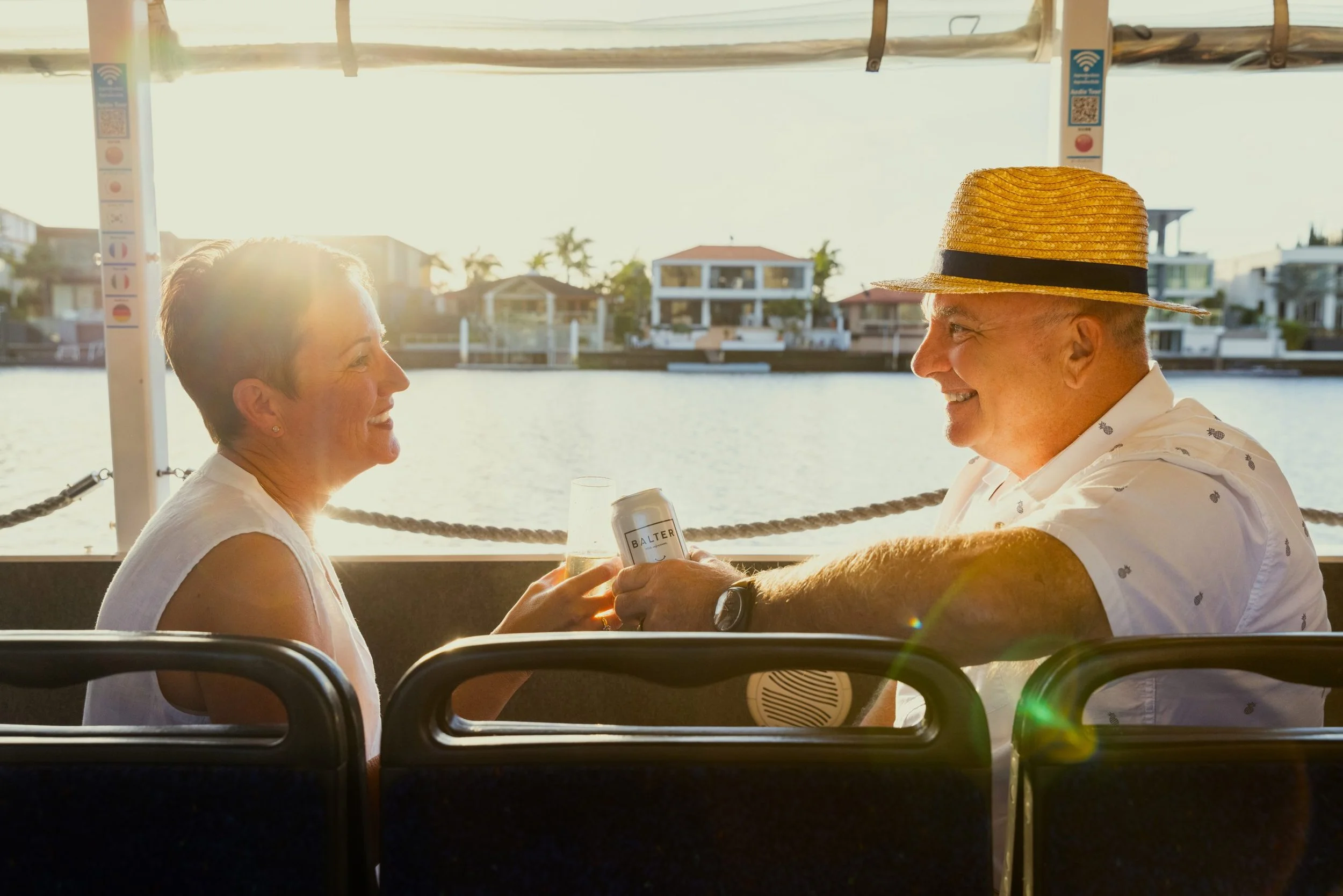 A man and woman enjoying drinks on a boat at sunset, with a waterway and modern houses in the background.