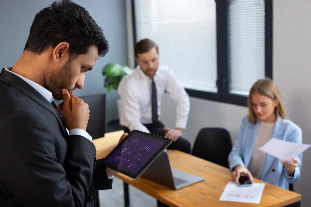 Business professionals in a meeting room, one man in a black suit reviewing data on a tablet, a woman holding papers and a phone, and another man sitting at the table looking at the tablet.